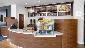 Zona de recepción con mostrador curvo de madera, máquina de café, vitrina refrigerada con aperitivos y un panel informativo.