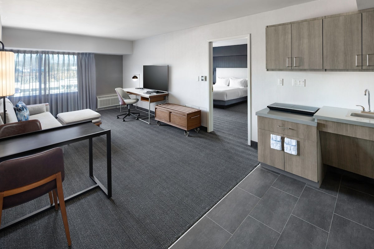 Quarto de hotel moderno com escrivaninha, cadeira e cozinha compacta.