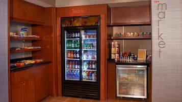 Un petit espace aménagé en épicerie, avec un réfrigérateur rempli de boissons et d’en-cas, des étagères garnies de produits emballés et un présentoir où sont exposés des produits alimentaires.