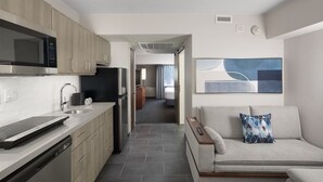 Cocina moderna equipada con lavavajillas, microondas y refrigerador. Hay un sofá con un cojín y un control remoto sobre una mesita de madera. Un pasillo conduce a un dormitorio con cama y ventana.