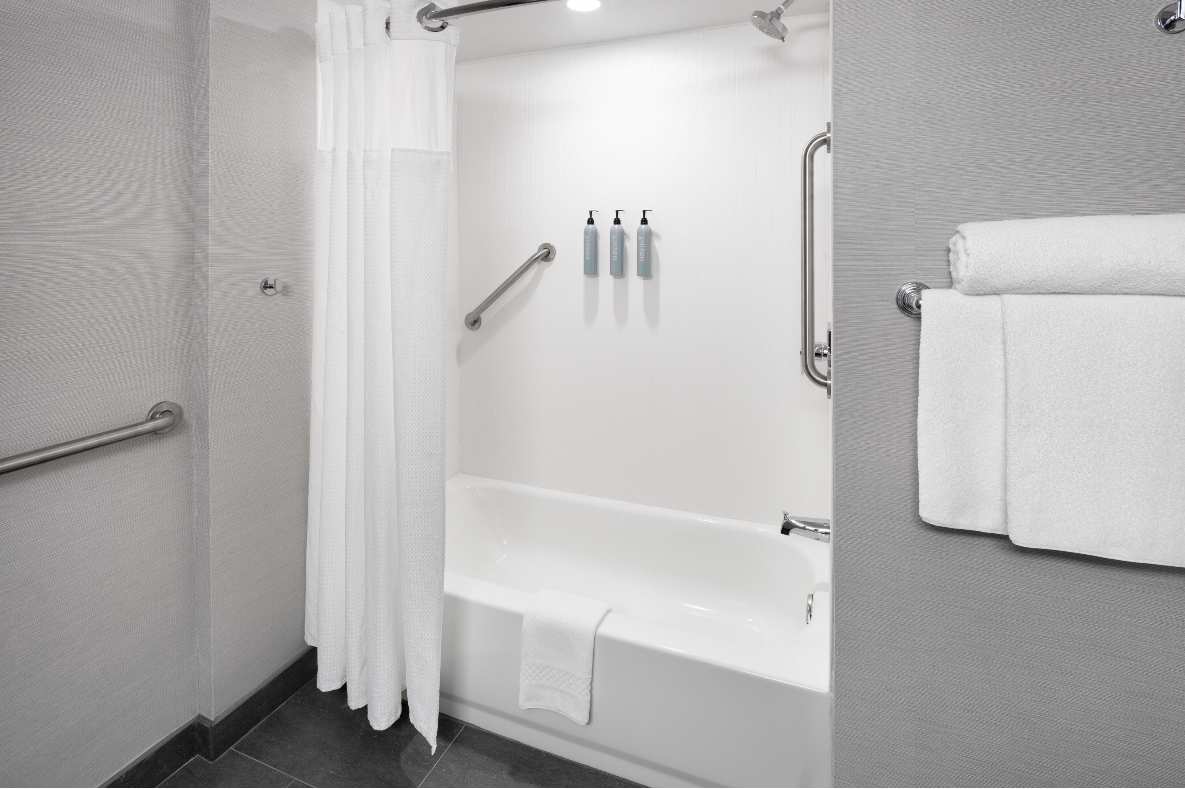 A modern bathroom with a bathtub, a shower area, and a handrail. There are three bottles of shampoo and a towel rack with a folded towel.