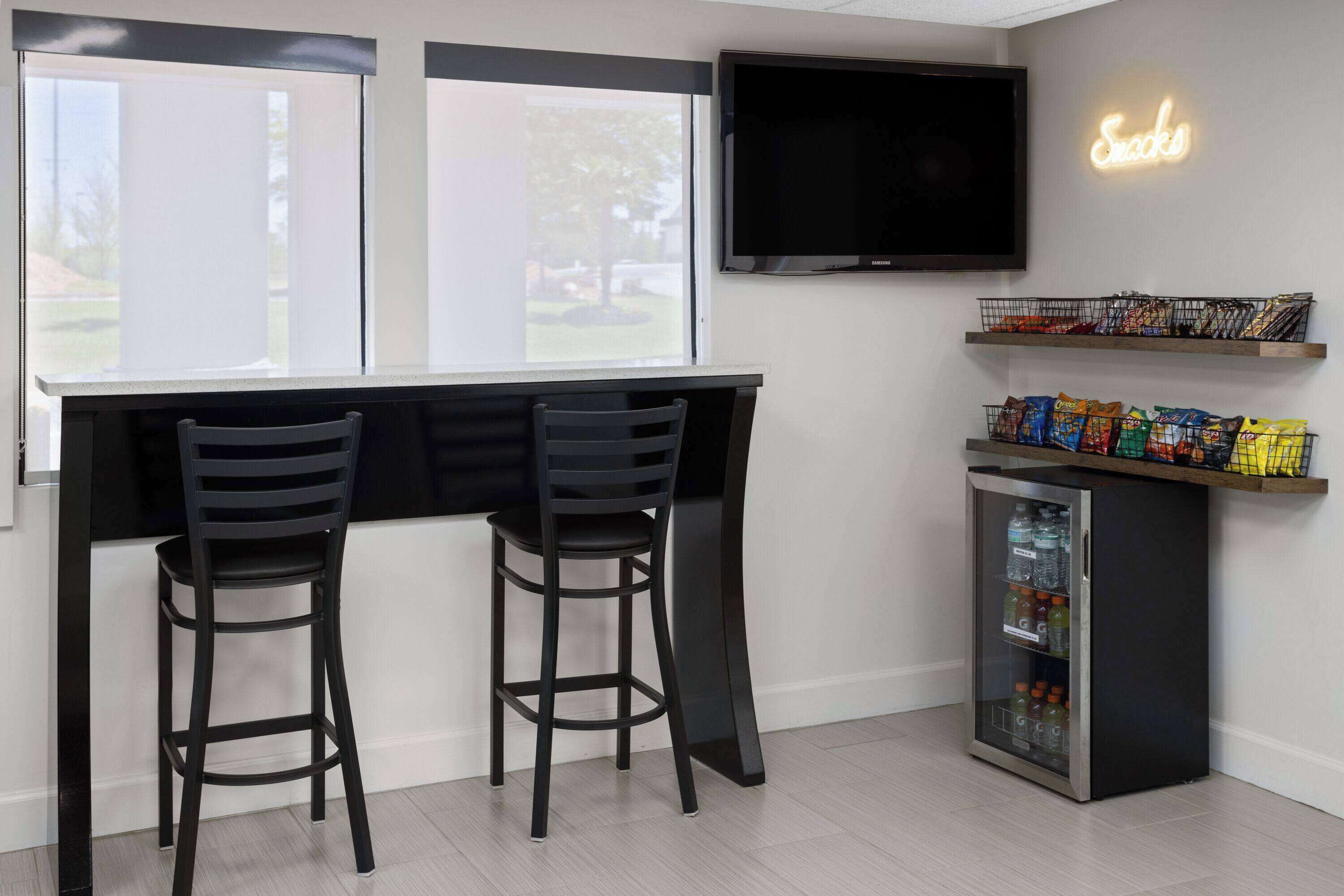 A modern kitchen with two black bar stools, a built-in TV, a neon sign, snack shelves, and a mini-fridge.