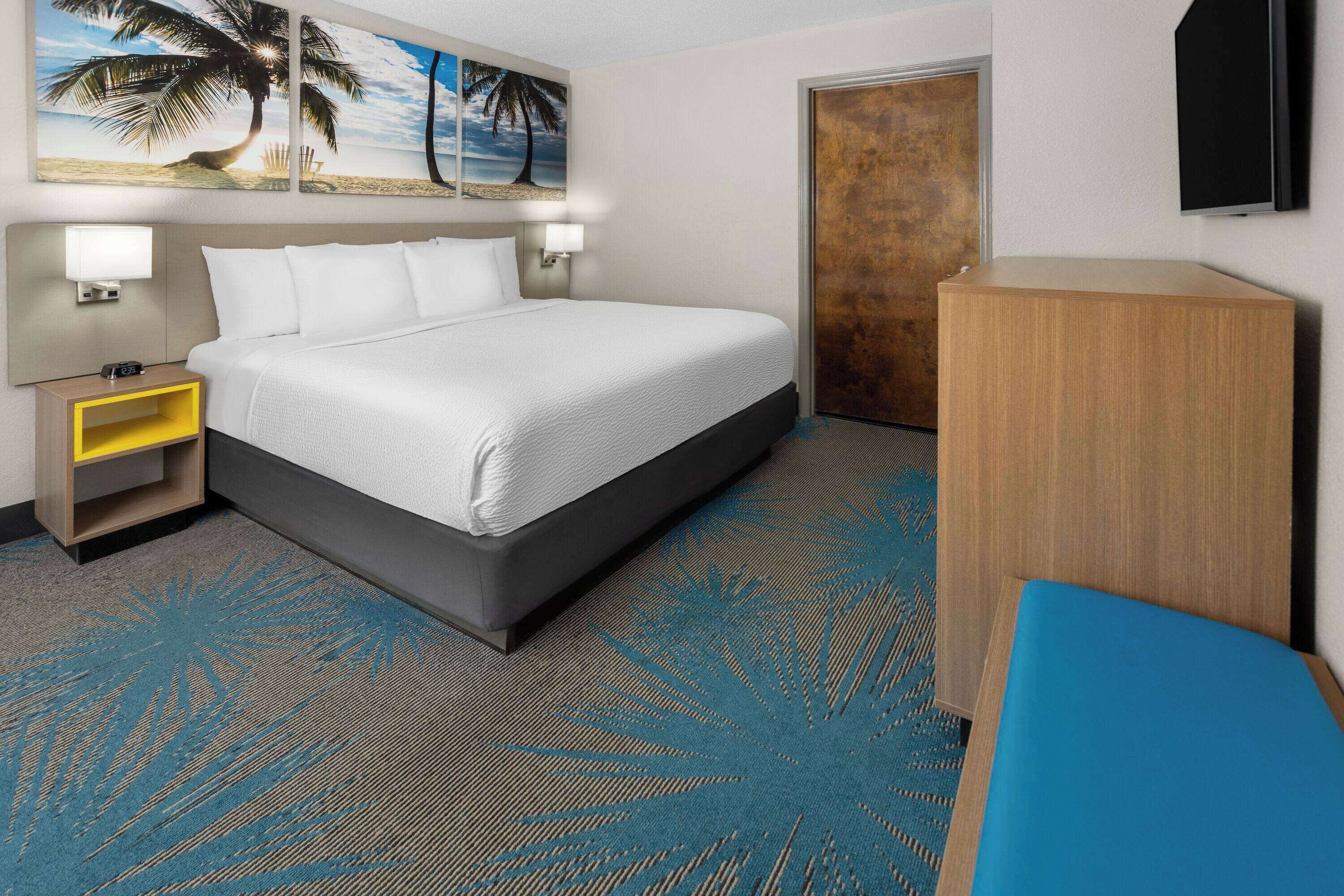 A hotel room with a bed, nightstand, lamp, and a television on the wall.
