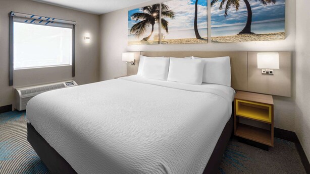 A hotel room with a large bed, a window with blinds, two wall-mounted lamps, a nightstand with a lamp, and a beach-themed painting above the bed. - Days Inn by Wyndham Acworth (Acworth)