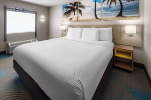 Une chambre d’hôtel avec un grand lit, une fenêtre avec des stores, deux lampes fixées au mur, une table de chevet avec une lampe et un tableau sur le thème de la plage au-dessus du lit.