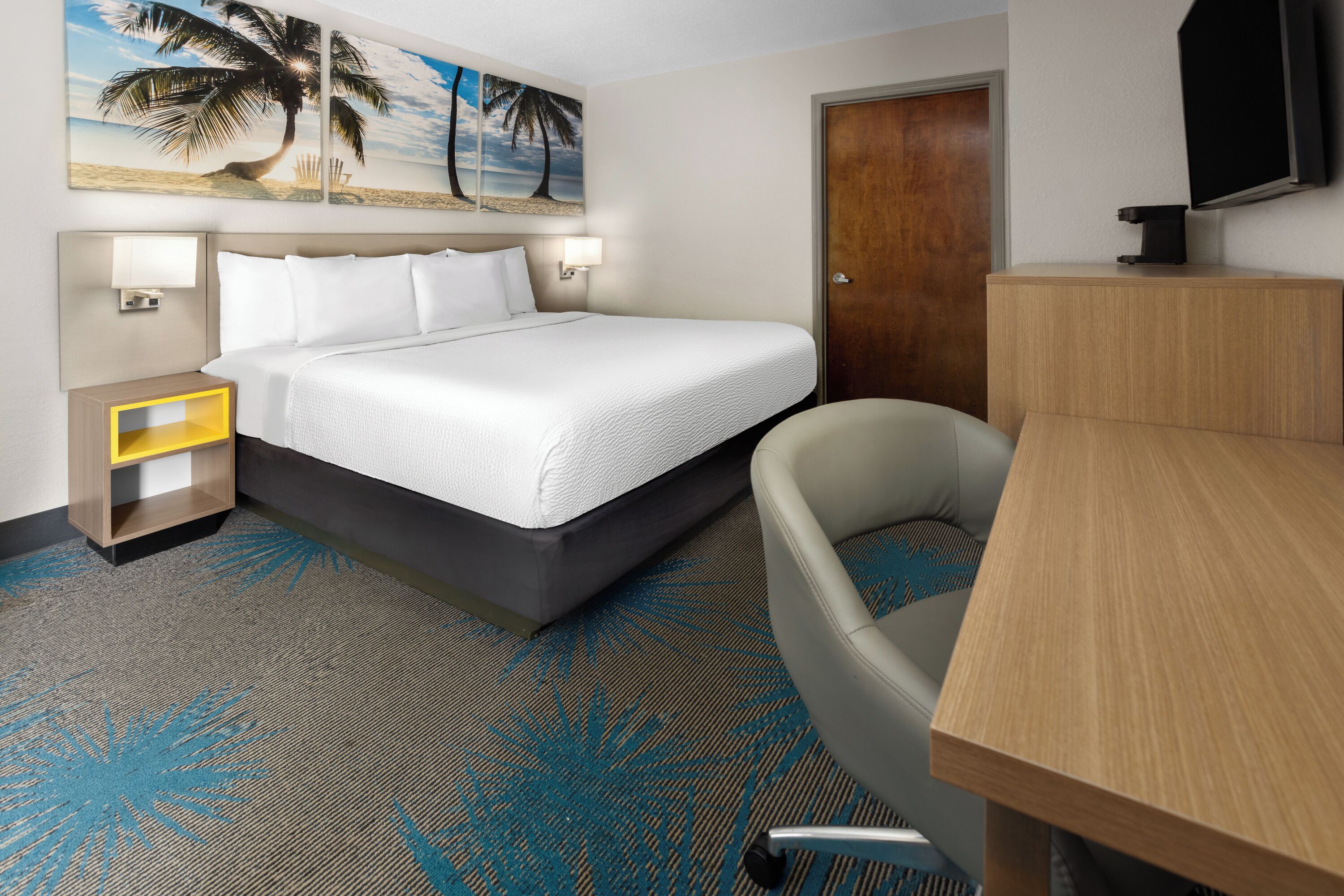 A hotel room with a bed, a desk, a chair, a nightstand, a TV, and a door.