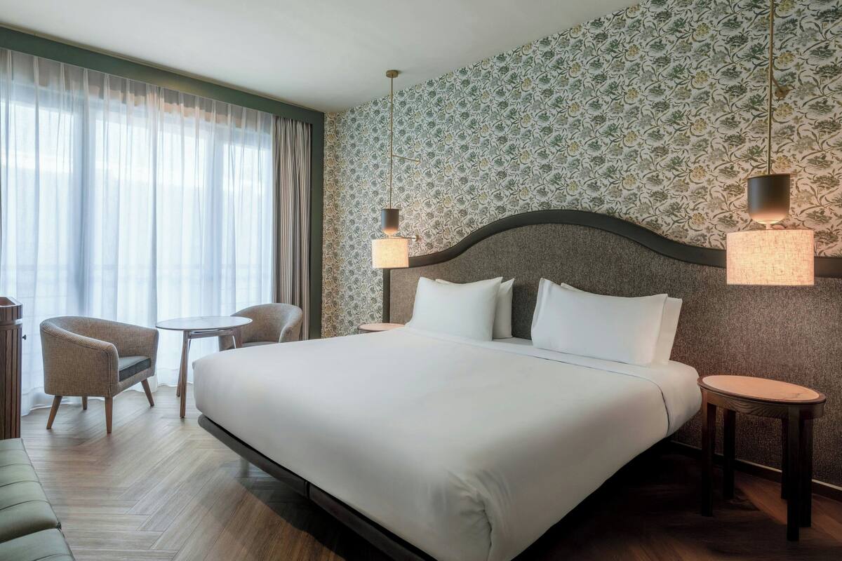 Een hotelkamer met een groot bed, twee nachtkastjes, een stoel, een klein tafeltje en behang met een patroon.
