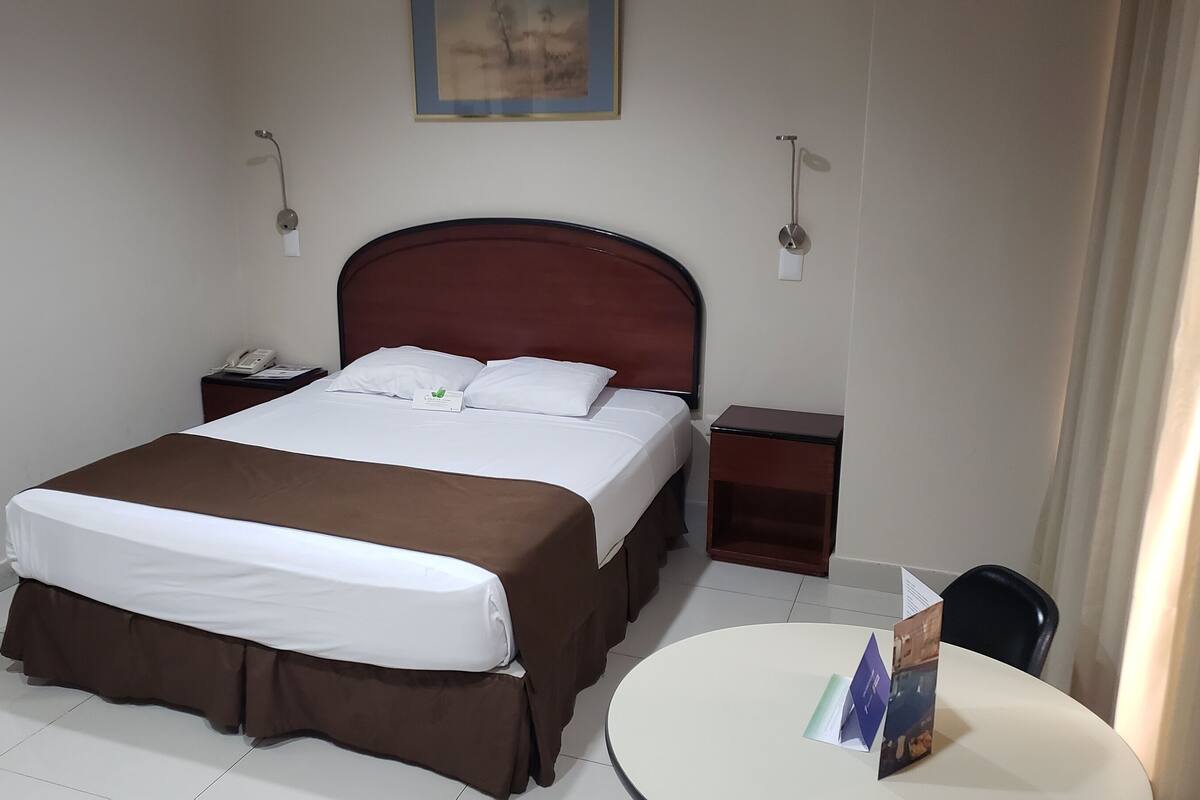 A hotel room with a bed, a bedside table with a phone, a small table with brochures, and a chair.