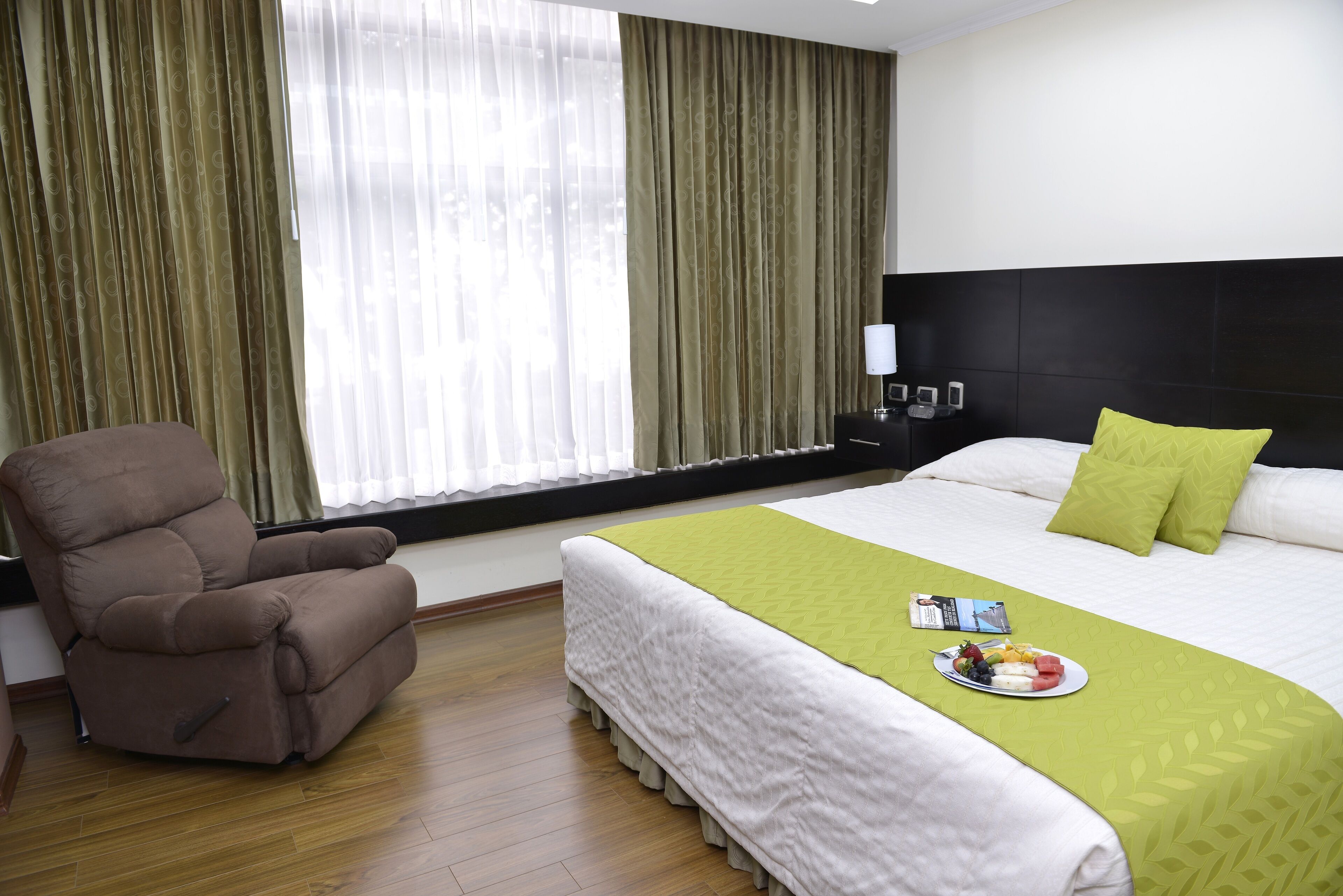 Habitación de hotel con cama, sillón reclinable, mesita de noche con lámpara y ventana con cortinas.