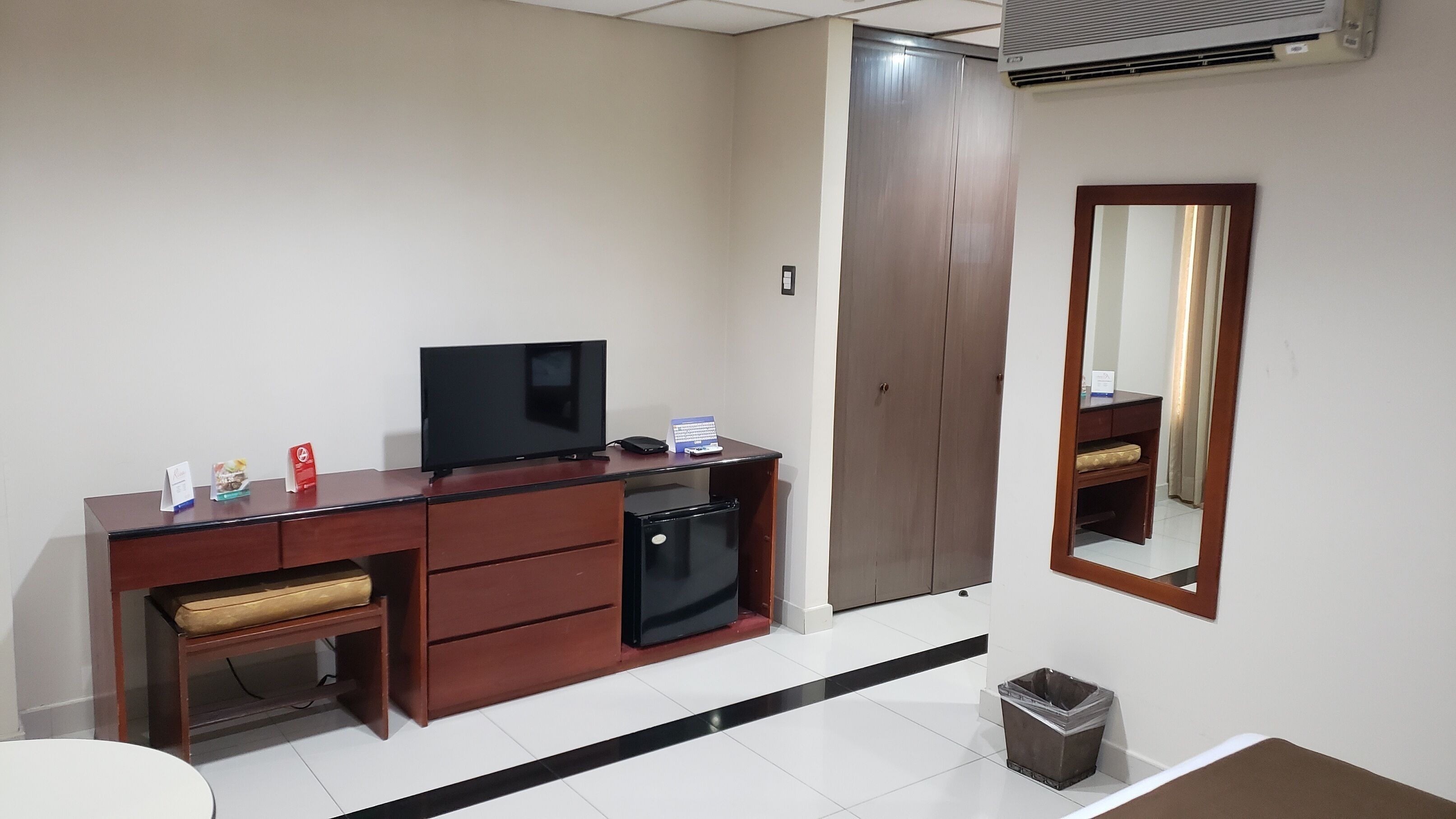 A hotel room with a wooden desk, a television, a small refrigerator, a trash bin, and a wardrobe.