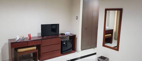 A hotel room with a wooden desk, a television, a small refrigerator, a trash bin, and a wardrobe.