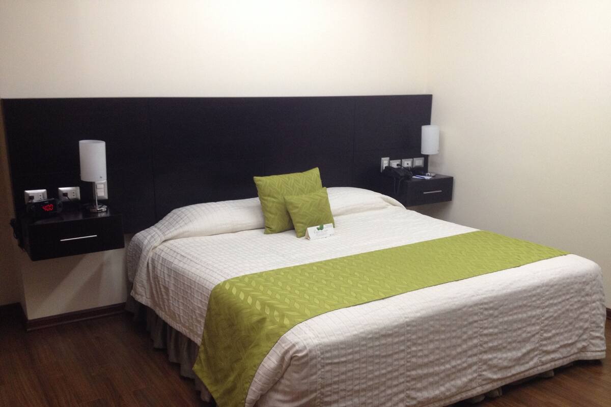 A neatly made bed with a green and white bedspread, a pillow, and a bedside table with a lamp and a clock.
