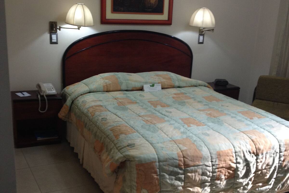 A hotel room with a bed, two bedside lamps, a nightstand with a phone, and a framed artwork on the wall.