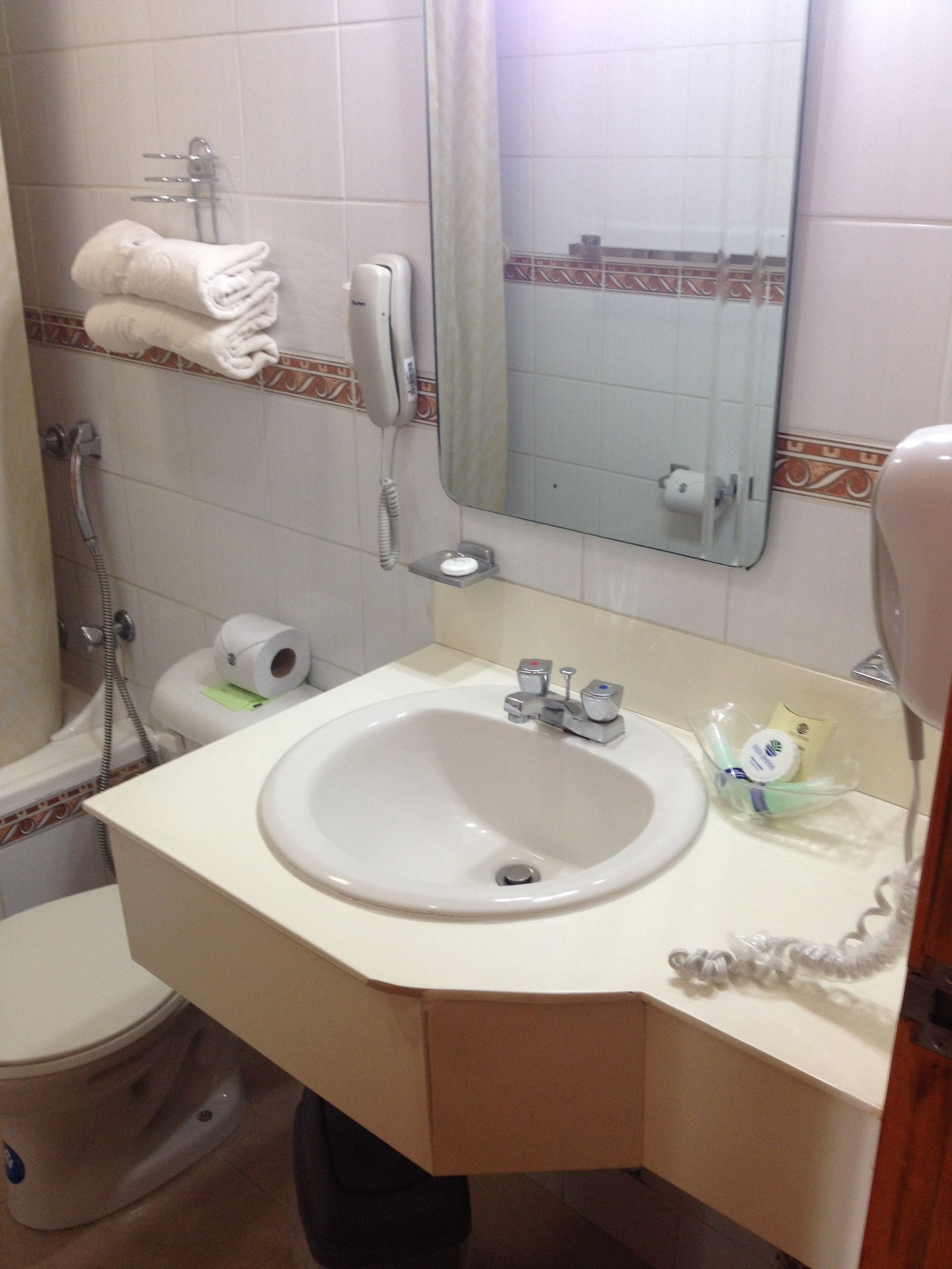 A bathroom with a sink, toilet, and shower. There is a towel rack with towels, a telephone, and a mirror.