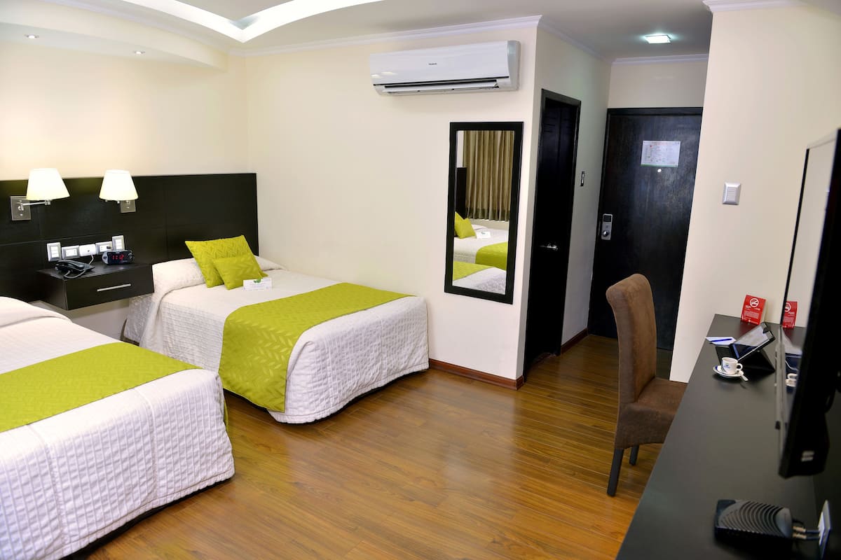 A hotel room with two beds, a desk, a chair, an air conditioner, and a mirror.