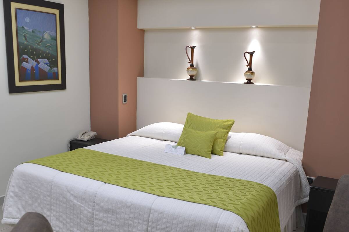 A hotel room with a bed, a desk with a laptop and a cup of coffee, and a painting on the wall.