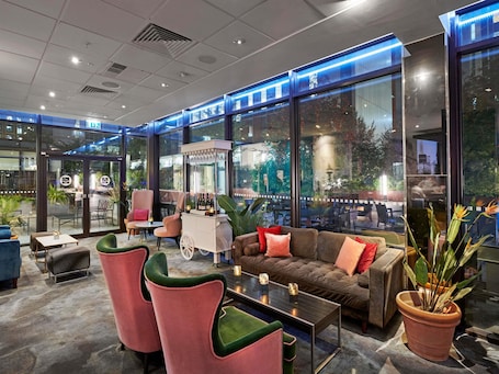 A modern lounge area with seating, a bar cart, and large windows offering views of the outdoors.. Novotel London West