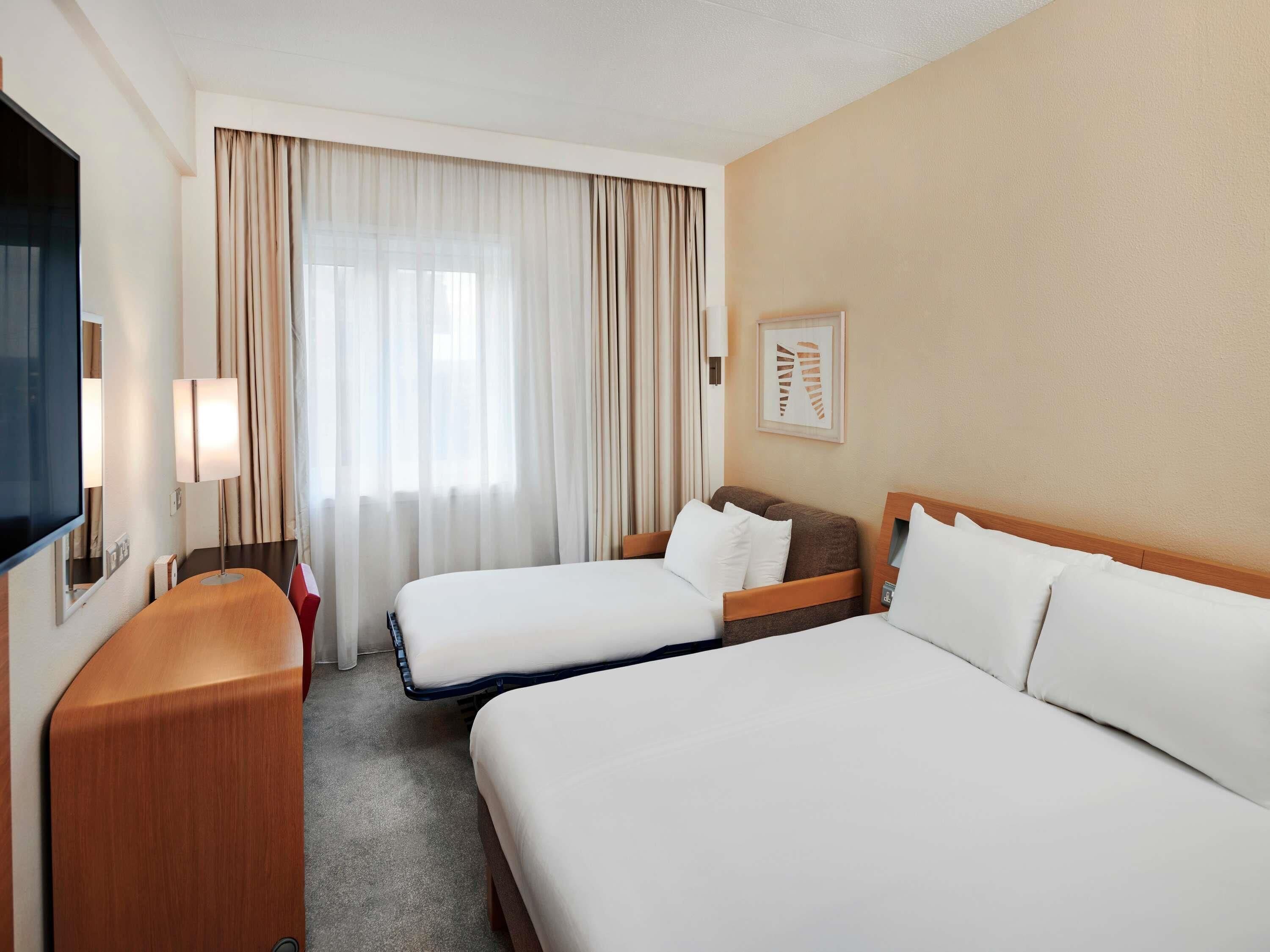 A hotel room with two beds, a TV, a desk, and a window with curtains.