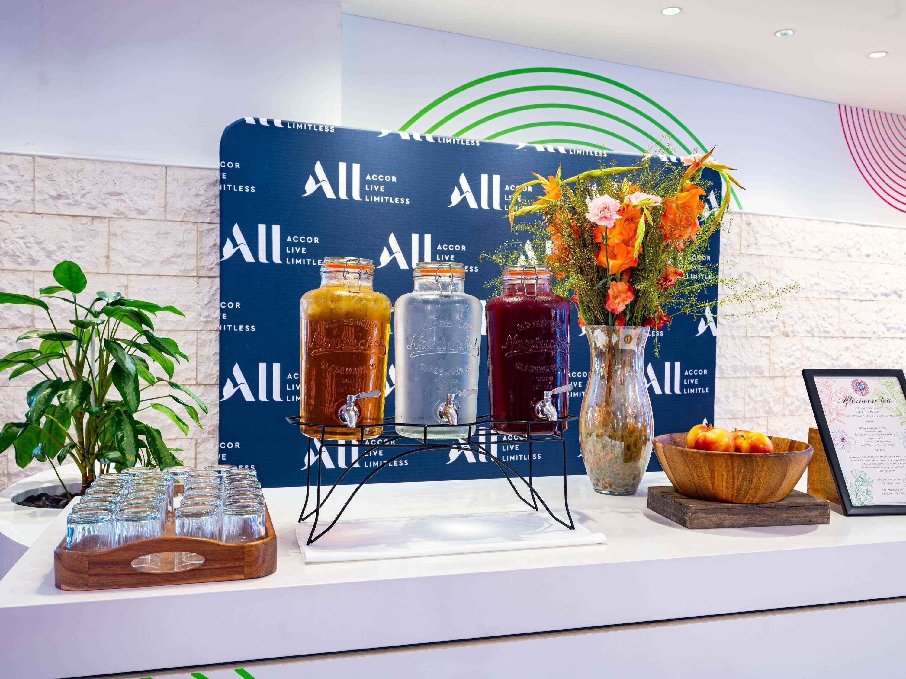 Zona de recepción con un fondo azul que muestra el texto 'All ACCOR LIVE LIMITLESS'. Hay tres dispensadores de vidrio con bebidas de diferentes colores, un jarrón con flores, una bandeja con vasos exprés y un menú enmarcado.