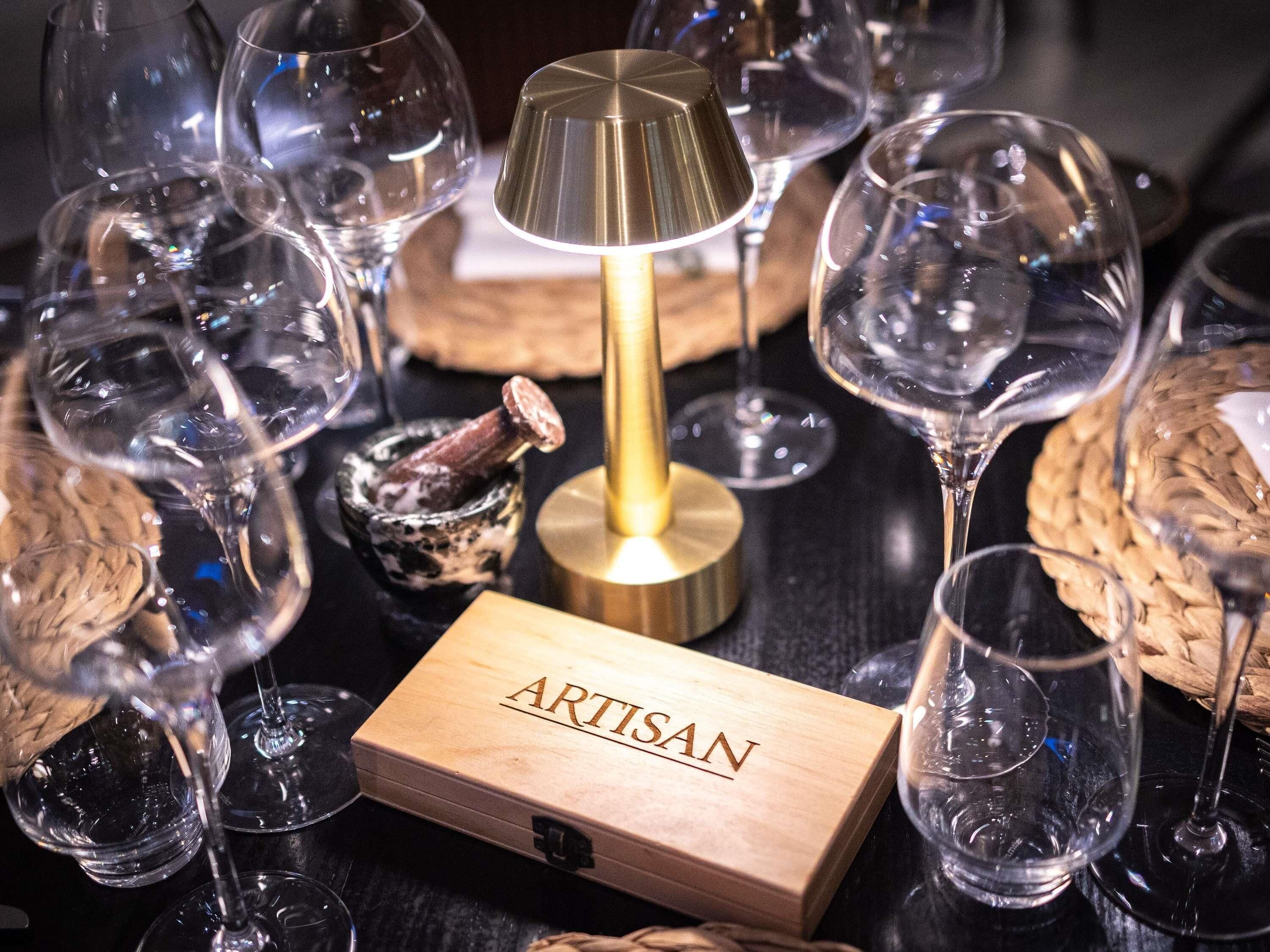 A collection of empty wine glasses, a wooden box labeled 'ARTISAN,' and a lamp on a dark surface.