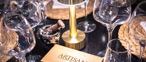 A collection of empty wine glasses, a wooden box labeled 'ARTISAN,' and a lamp on a dark surface.