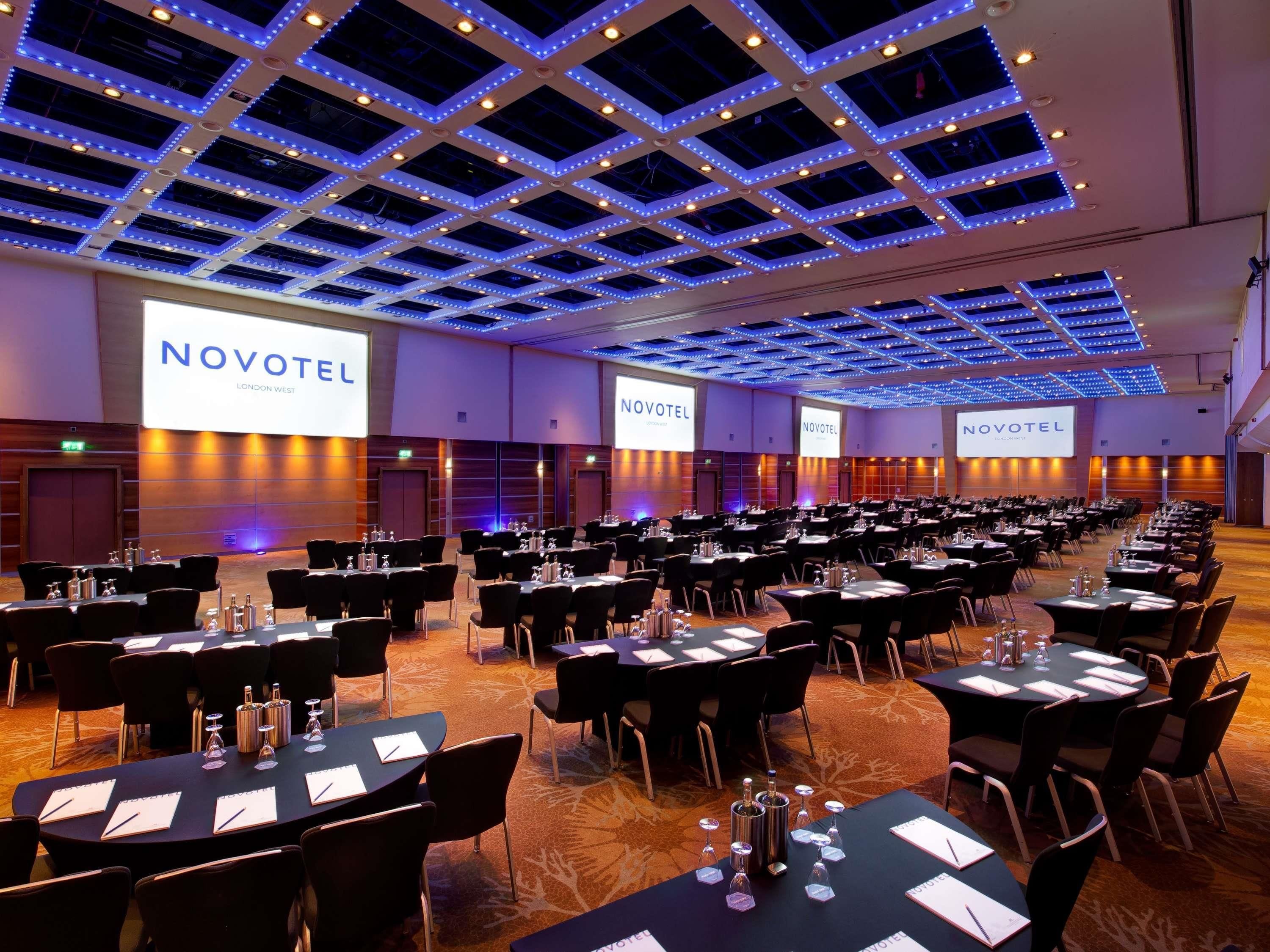 A conference room with multiple tables, chairs, and a large screen displaying 'NOVOTEL LONDON WEST'.