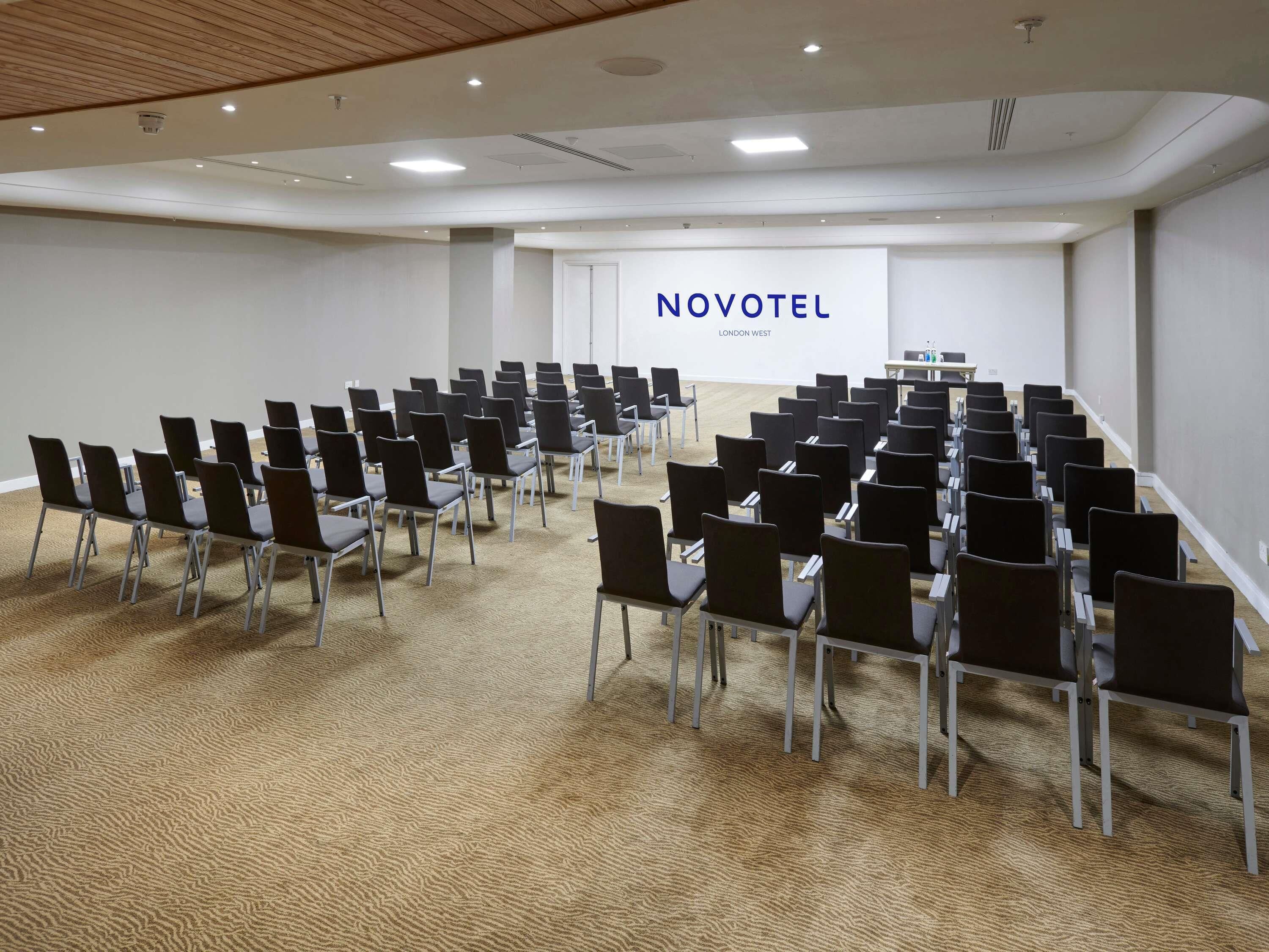 Un salón de conferencias con filas de sillas, un atril y una gran pantalla que muestra 'NOVOTEL LONDON WEST'.