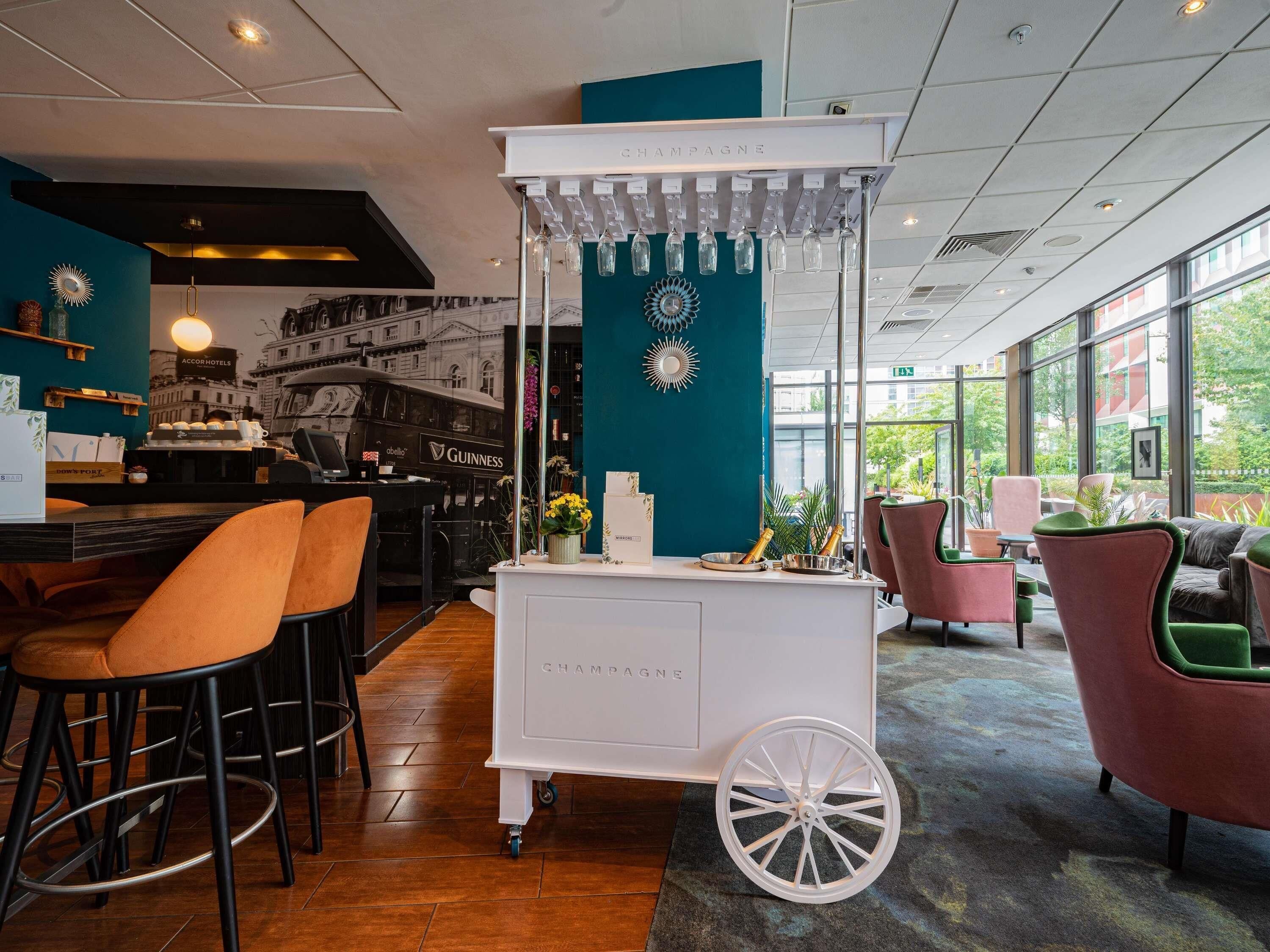 A bar area with a white cart labeled 'CHAMPAGNE,' bar stools, and a view of Accor Hotels through the window.