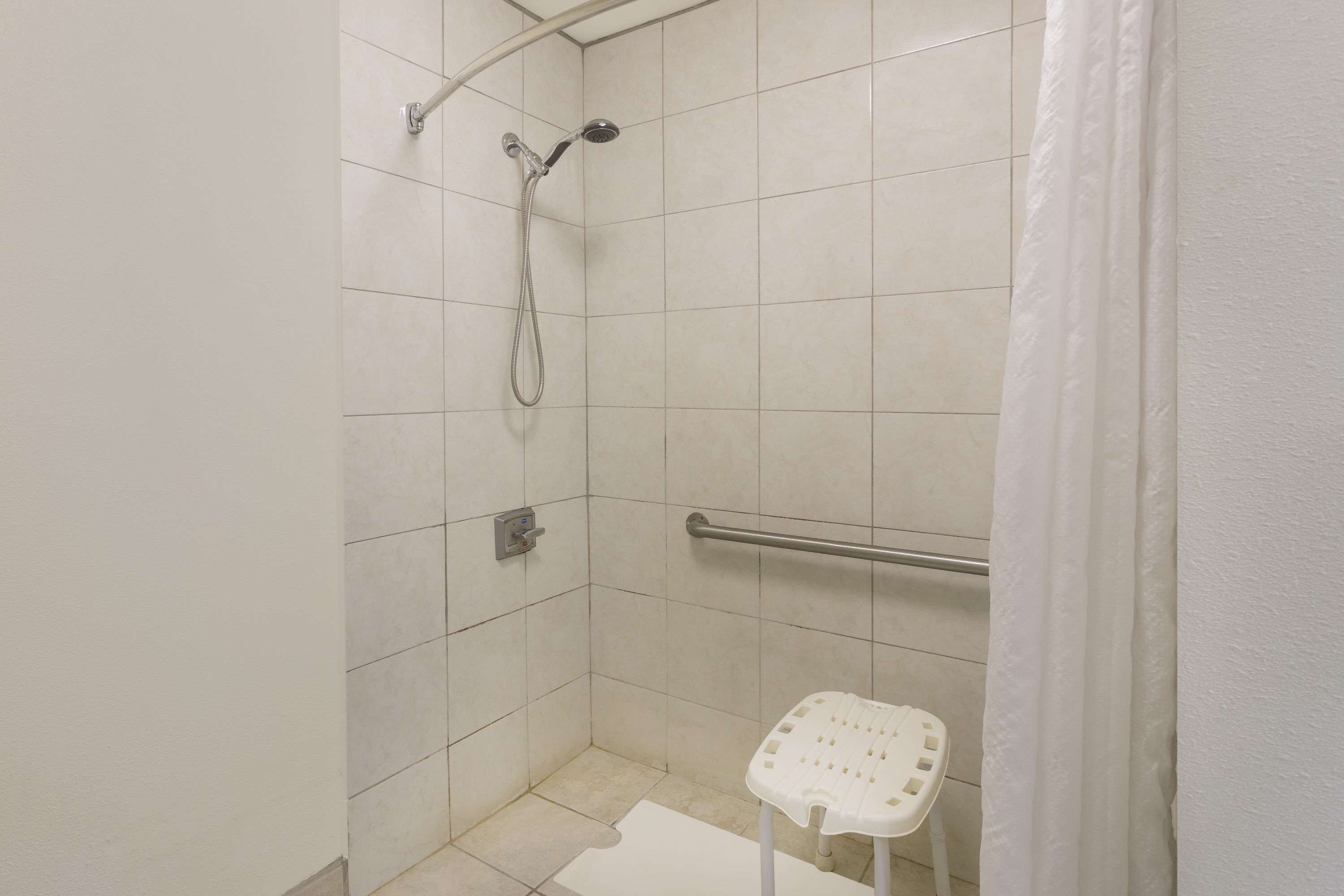 Pequeño box de ducha con azulejos, una barra de apoyo, un asiento blanco y una cortina blanca.