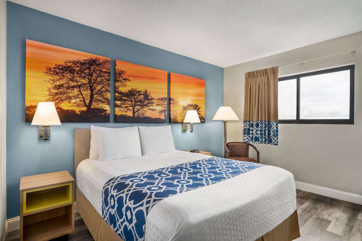 A hotel room with a bed, two wall-mounted lamps, a nightstand, a window with curtains, and a painting of a sunset.