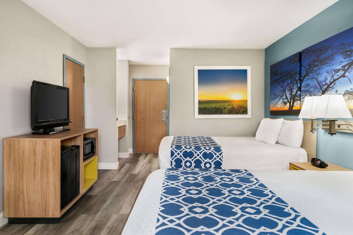 A hotel room with a bed, a TV on a wooden cabinet, a painting of a sunset, and a lamp on a bedside table.