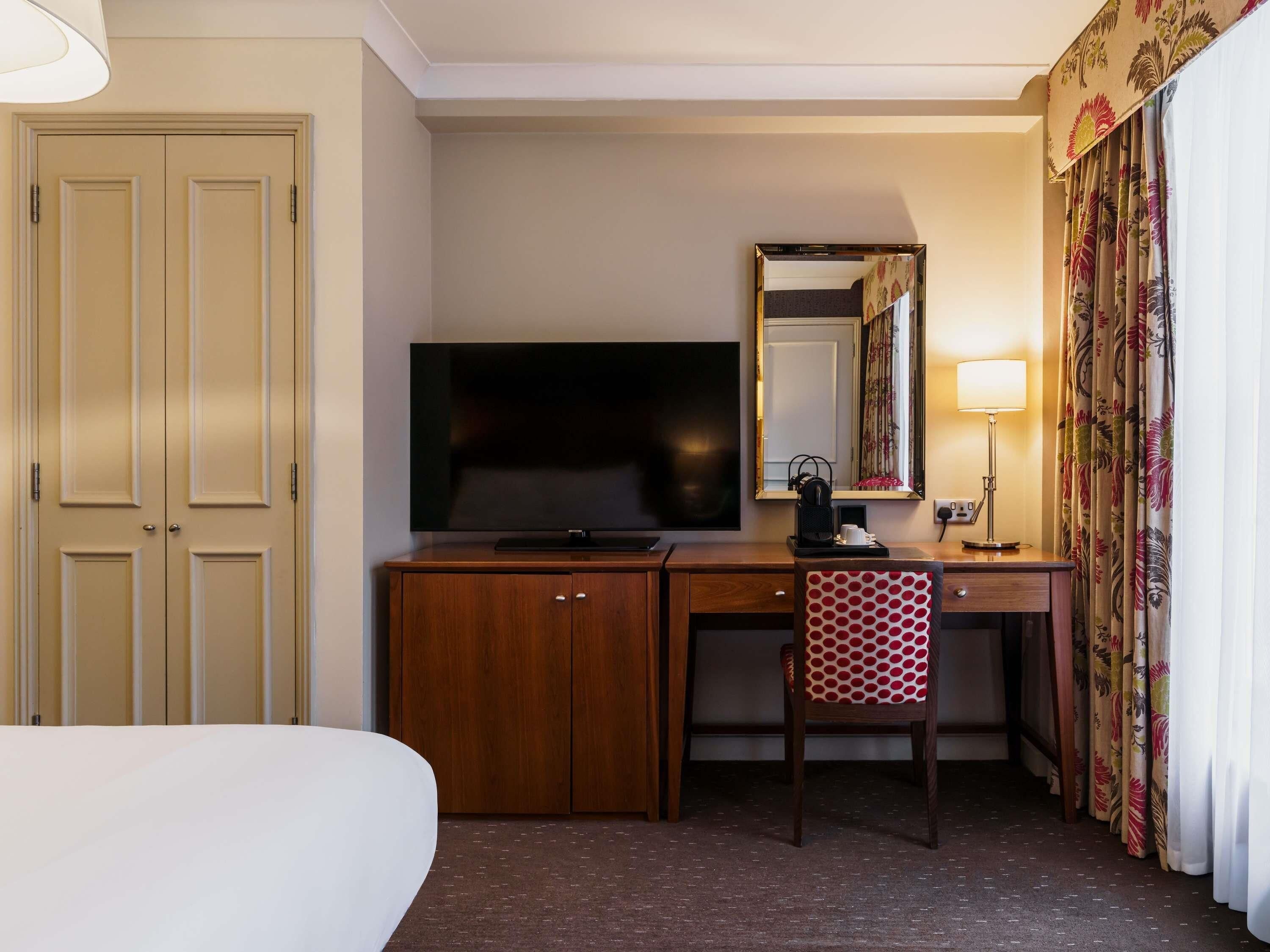 A hotel room with a bed, a desk with a television, a chair, a mirror, and curtains.