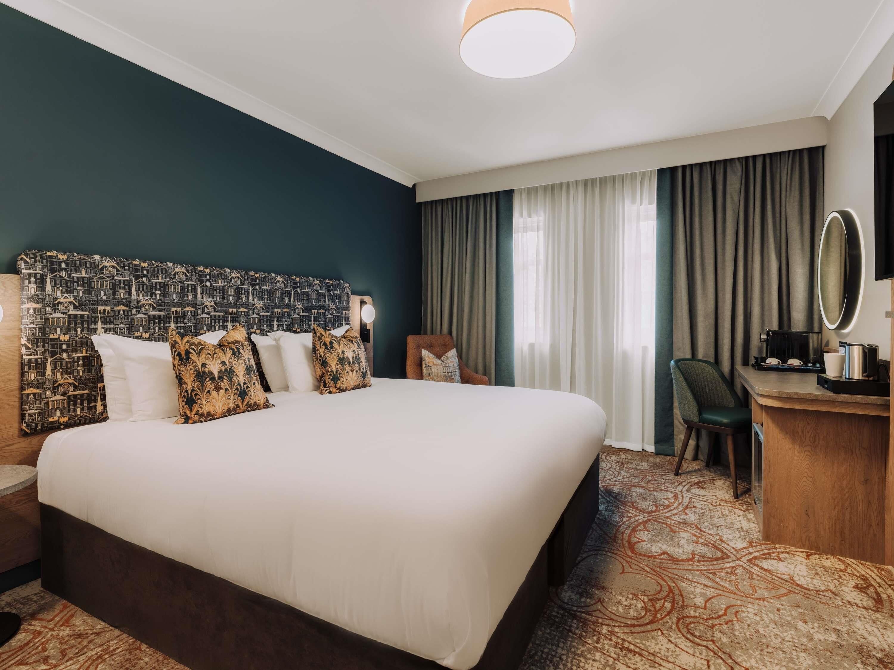 A hotel room with a large bed, a desk with a mirror, and a view of the city through the window.