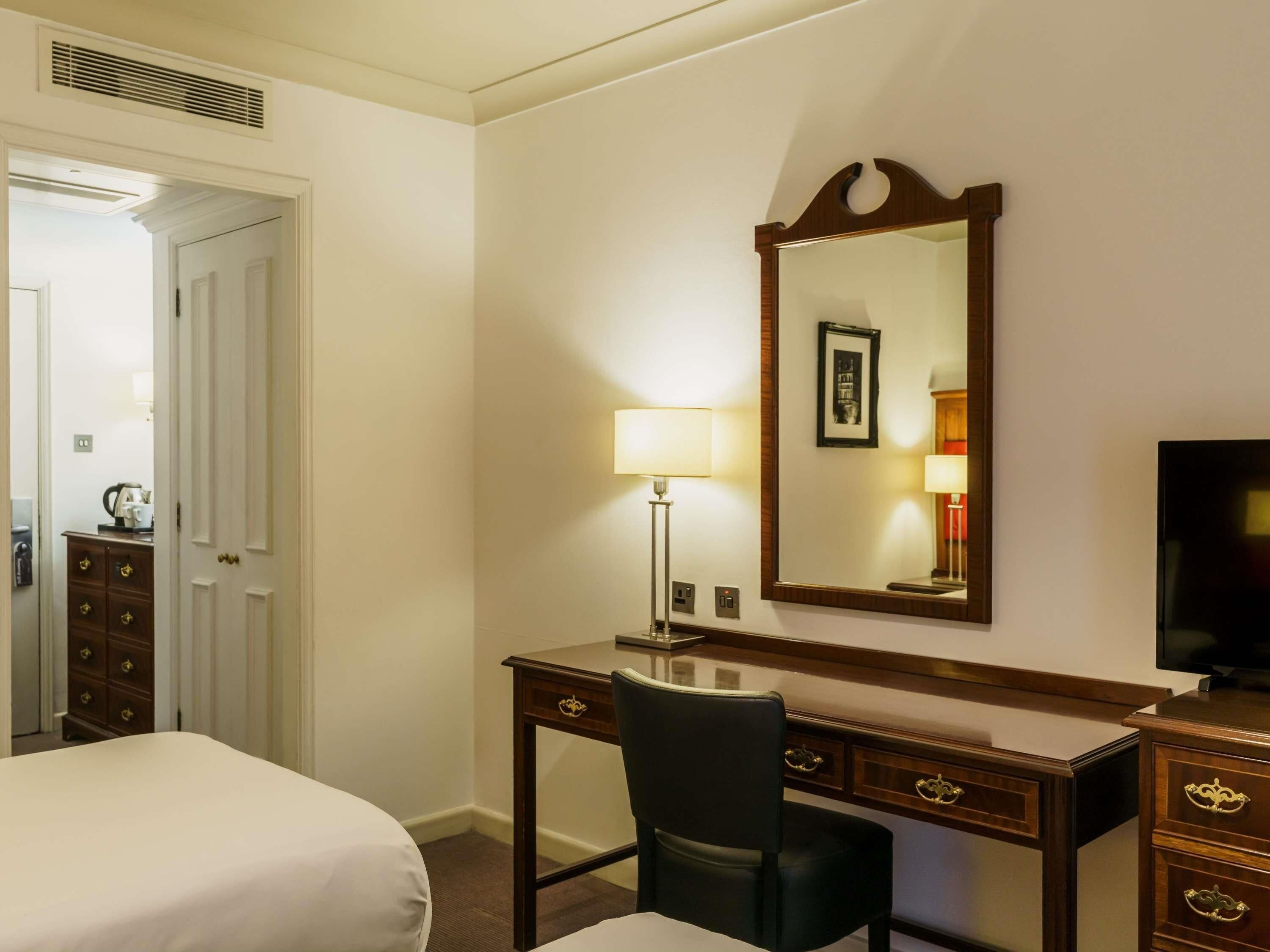Ein Hotelzimmer mit einem Bett, einem Schreibtisch mit Stuhl, einem Fernseher, einem Spiegel und einer Tür zu einem weiteren Raum.