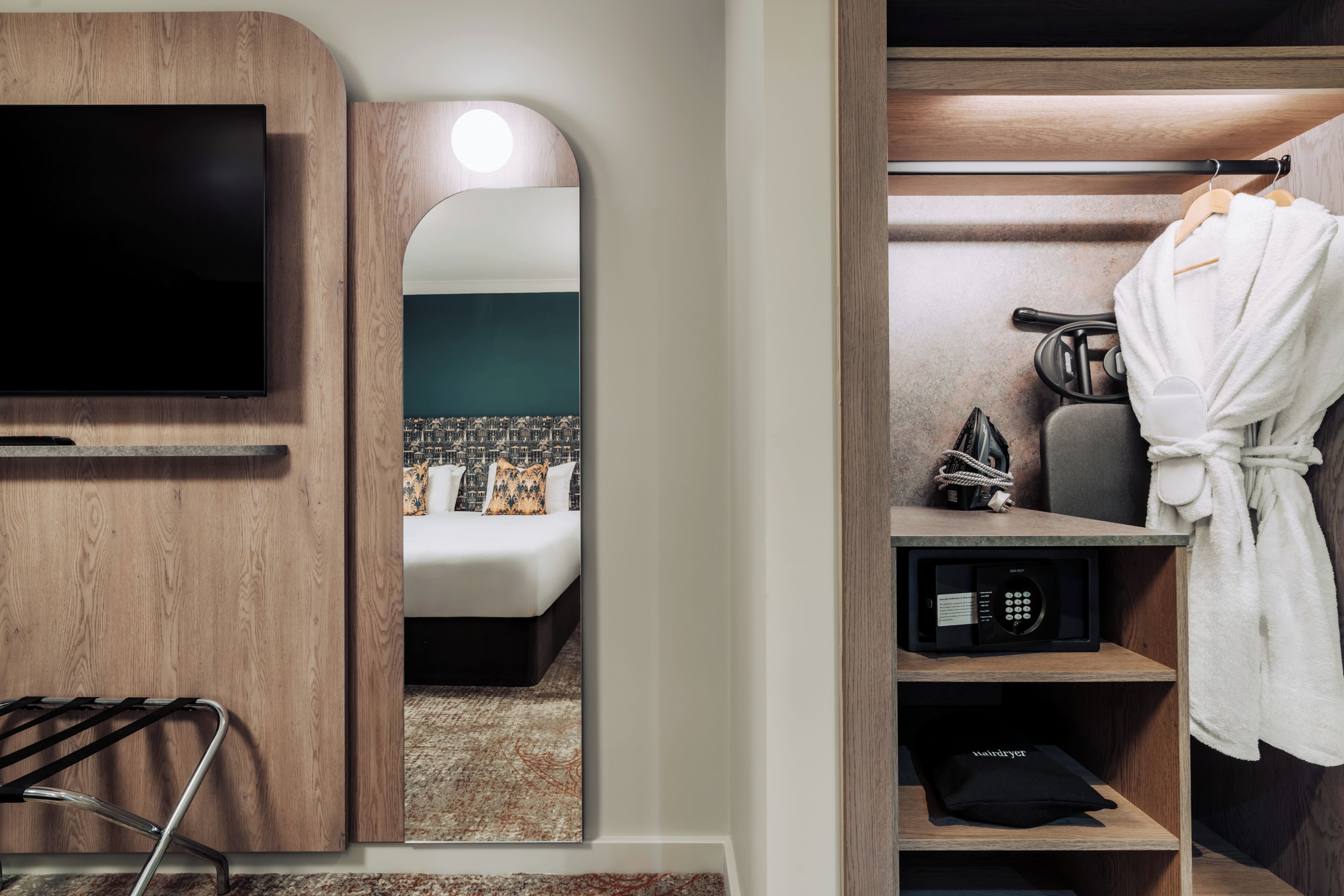 Habitación de hotel con televisión, espejo, bata, secador de pelo y estante con artículos de aseo.