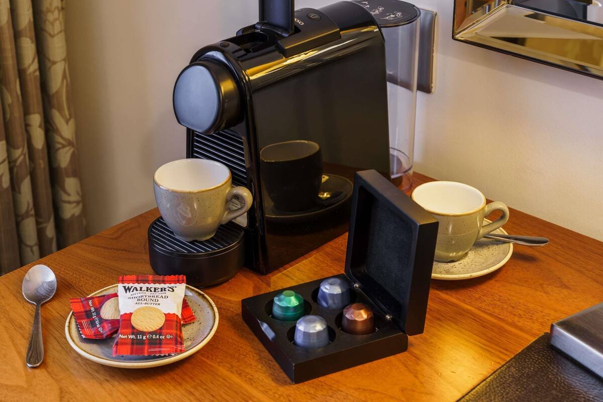 Una cafetera, dos tazas, un plato con galletas, una cuchara y una caja de galletas Walkers sobre una mesa de madera.