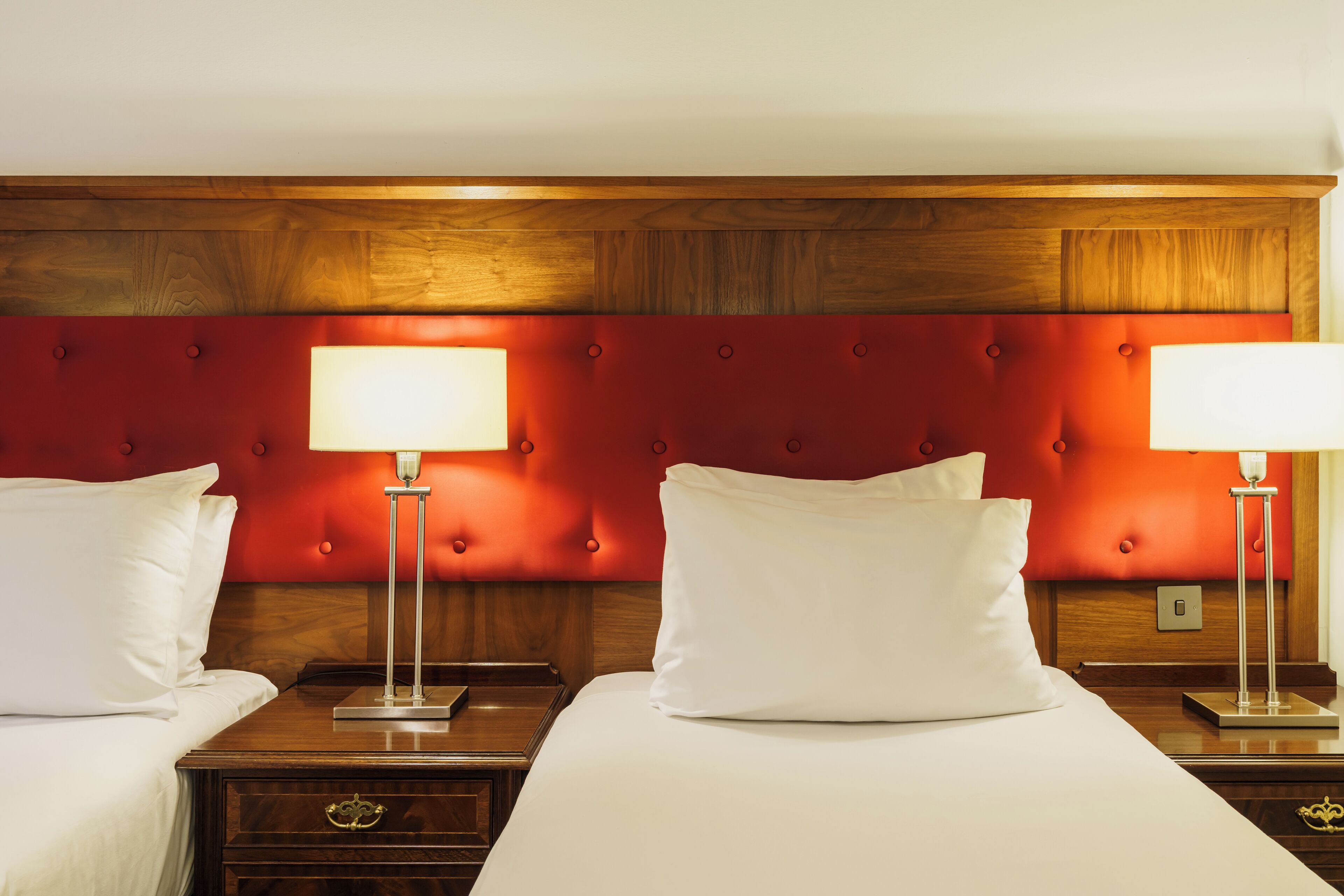 A hotel room with two beds, each with a wooden headboard and a red upholstered section. There are bedside tables with lamps and a nightstand with a drawer.