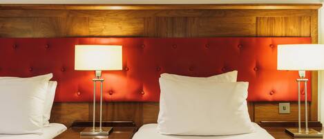 A hotel room with two beds, each with a wooden headboard and a red upholstered section. There are bedside tables with lamps and a nightstand with a drawer.