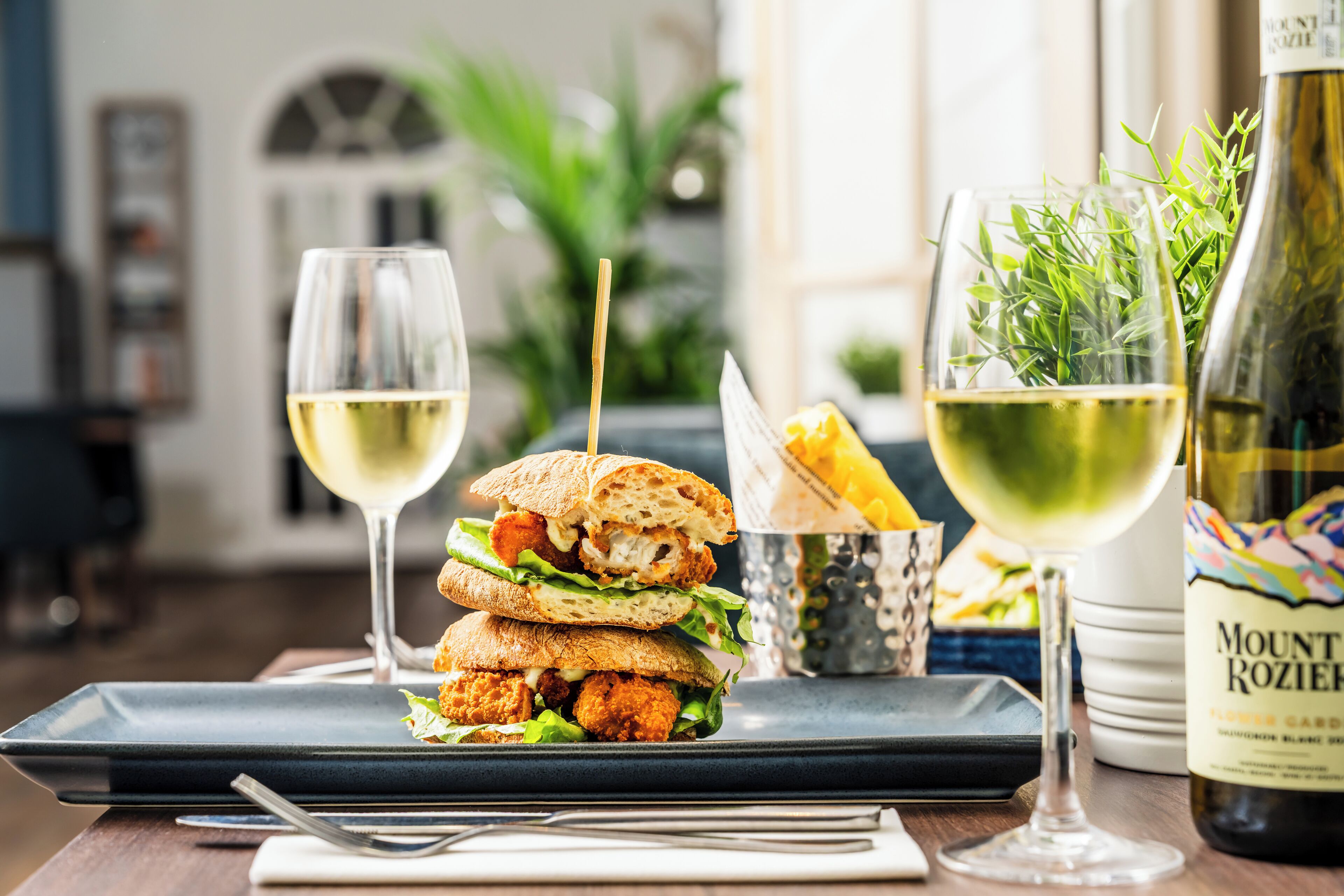 A sandwich with a toothpick, a glass of white wine, and a bottle of wine on a table.