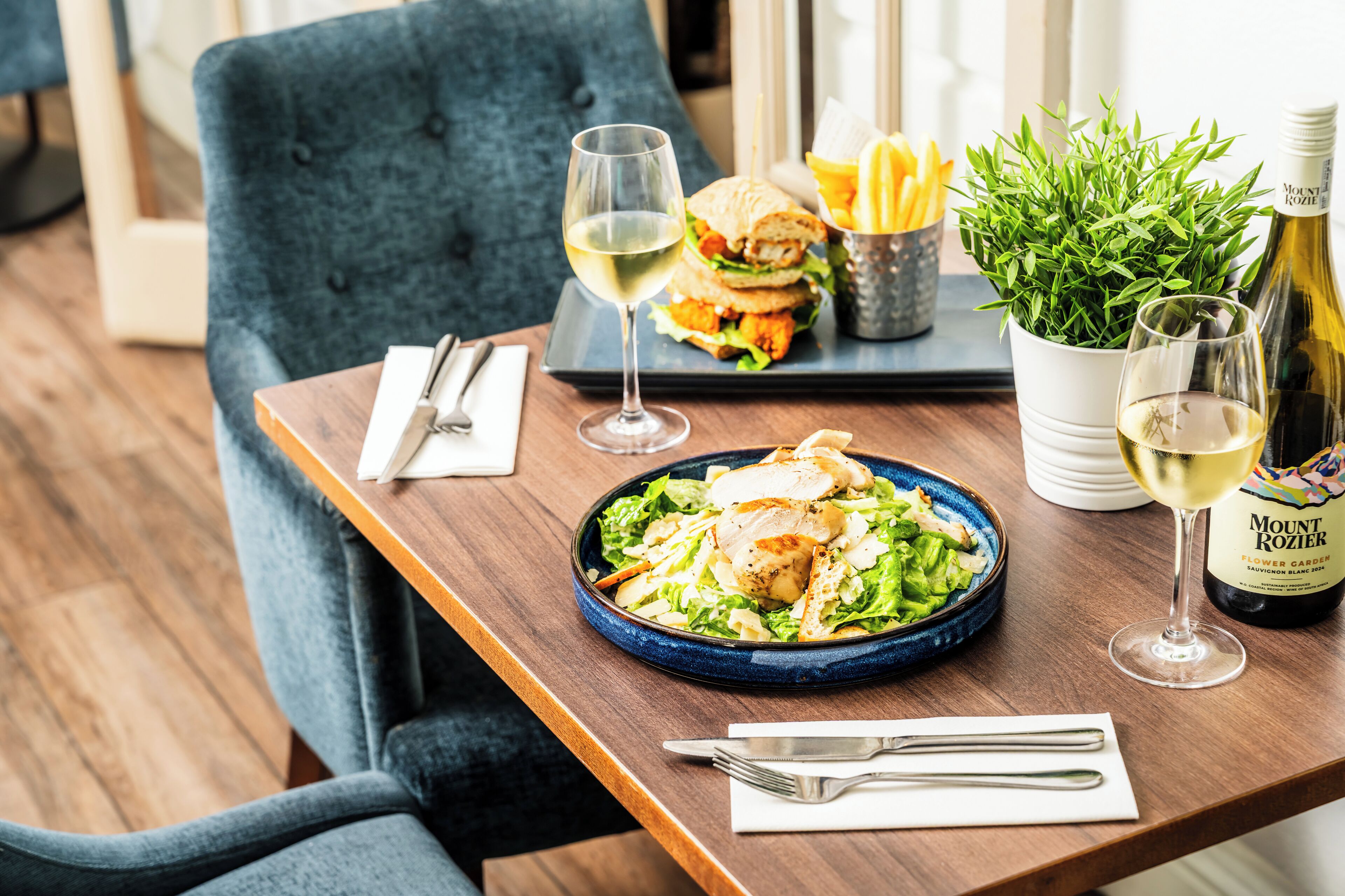 A dining table with a salad, a sandwich, fries, a bottle of wine, and a potted plant.