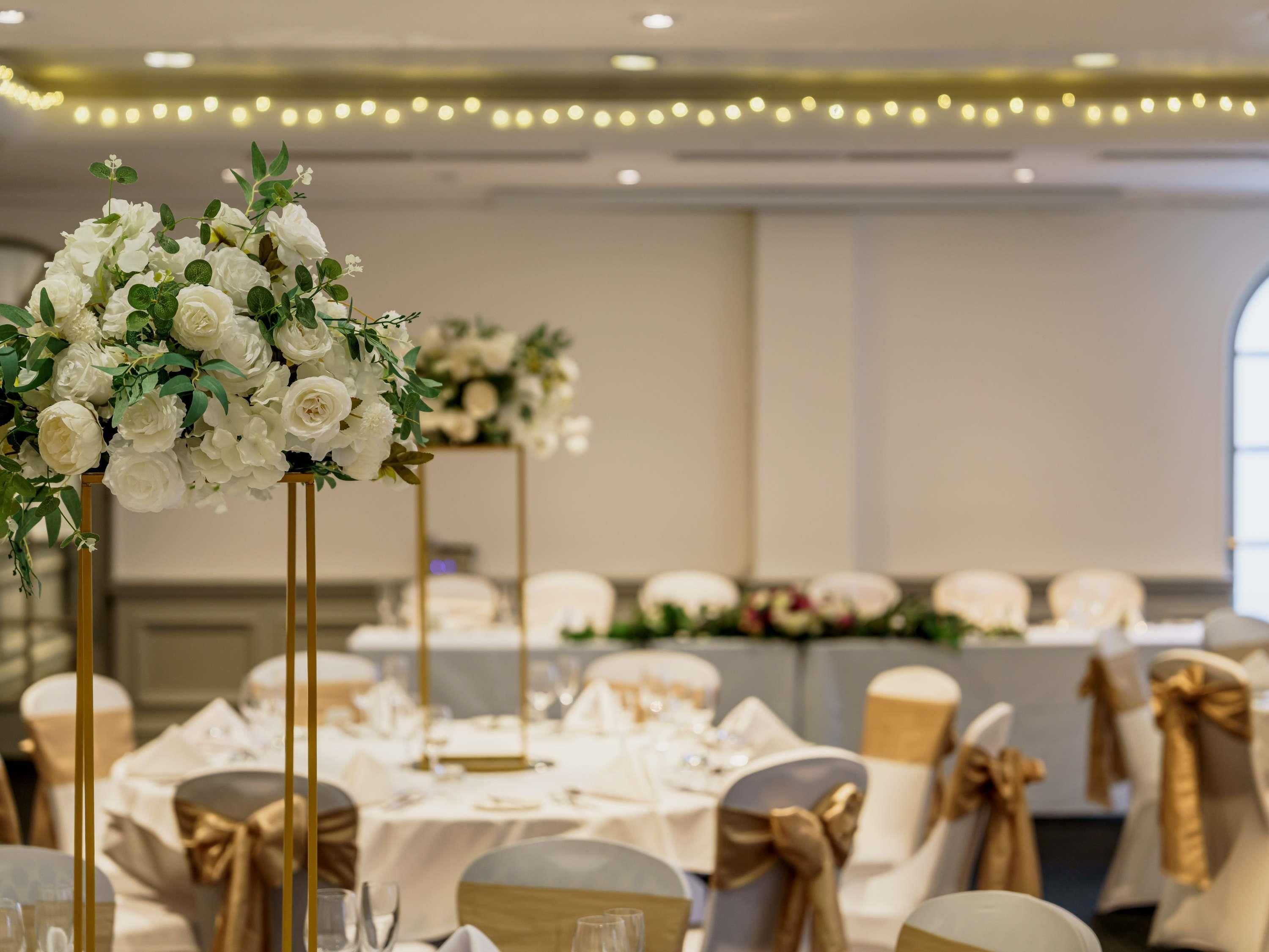 Une salle de banquet avec des tables rondes, des rubans dorés sur les chaises et des compositions florales en guise de décoration centrale.