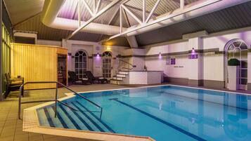 Una piscina interior con vista despejada al área de la piscina, una escalera que da acceso a ella y un área de descanso con sillas y una pequeña mesa.