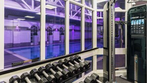 Un gimnasio con una hilera de mancuernas, una máquina de pesas y ventanas amplias con iluminación interior morada.