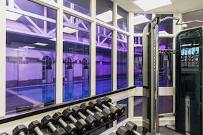 Un gimnasio con una hilera de mancuernas, una máquina de pesas y ventanas amplias con iluminación interior morada.