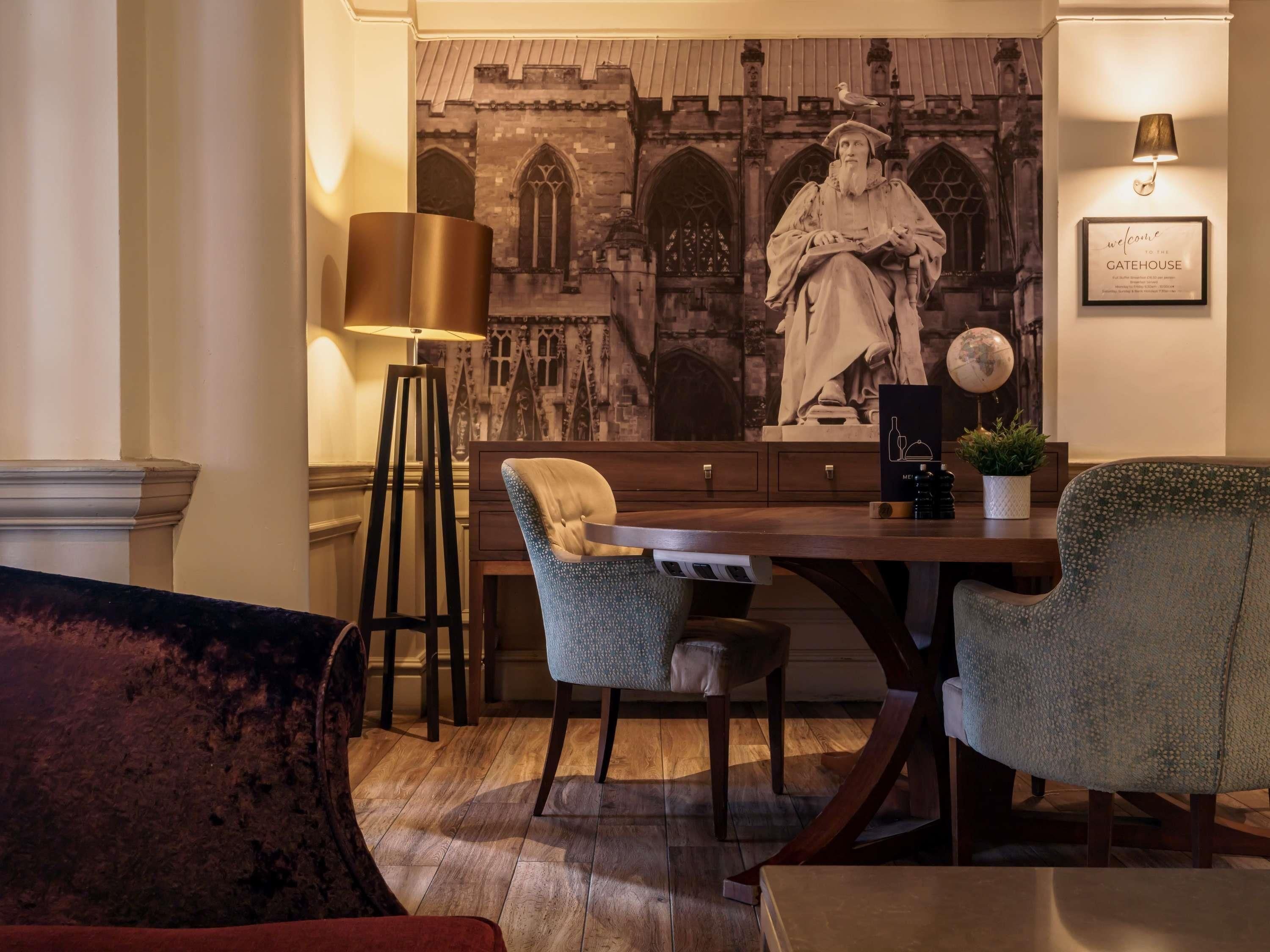 A dining area with a wooden table, two upholstered chairs, a tall floor lamp, and a wall mural of a building with a statue.