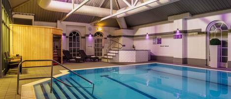 An indoor swimming pool with a clear view of the pool area, a set of stairs leading into the pool, and a lounge area with chairs and a small table.