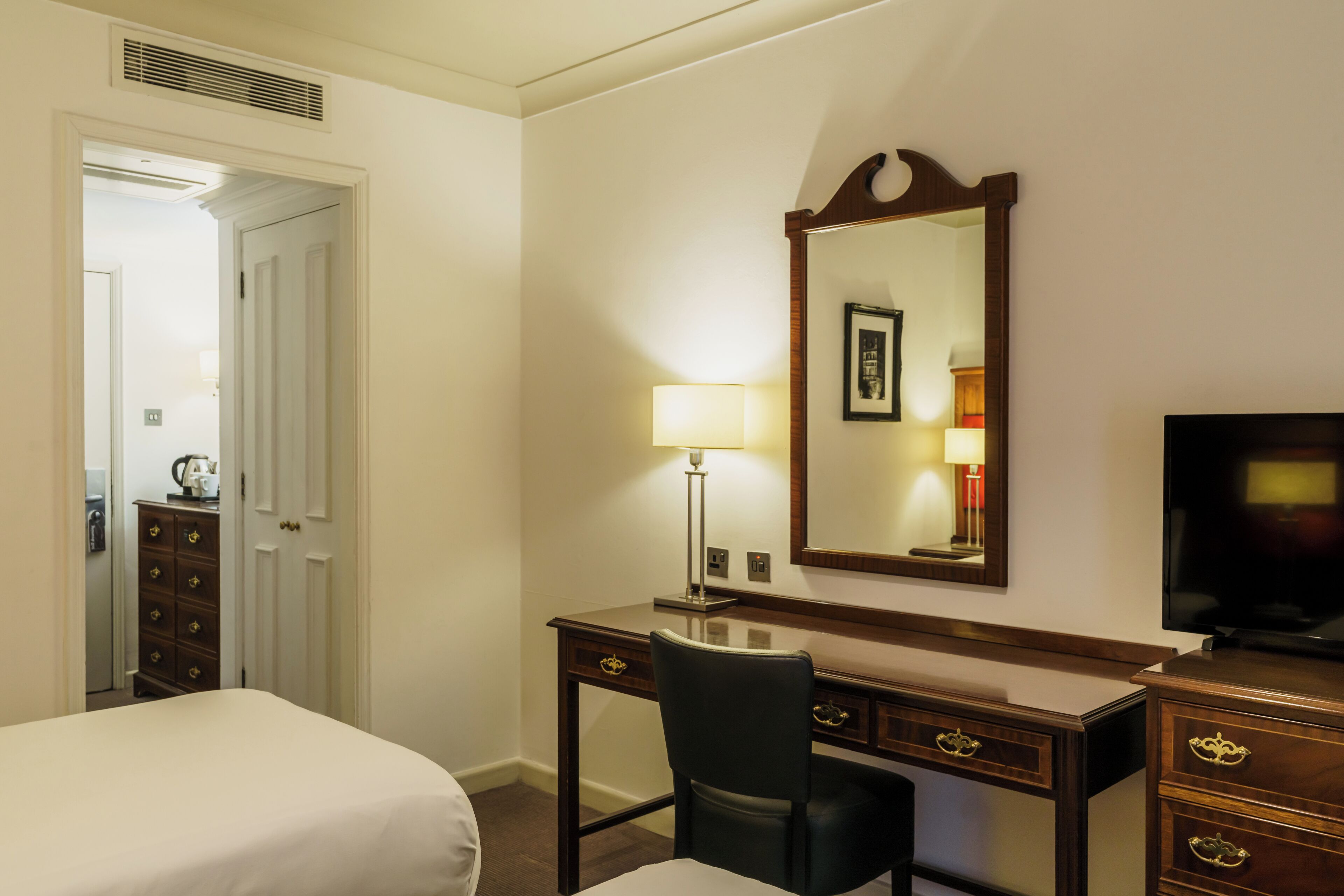 Habitación de hotel con cama, escritorio con silla, cómoda, televisor y espejo.