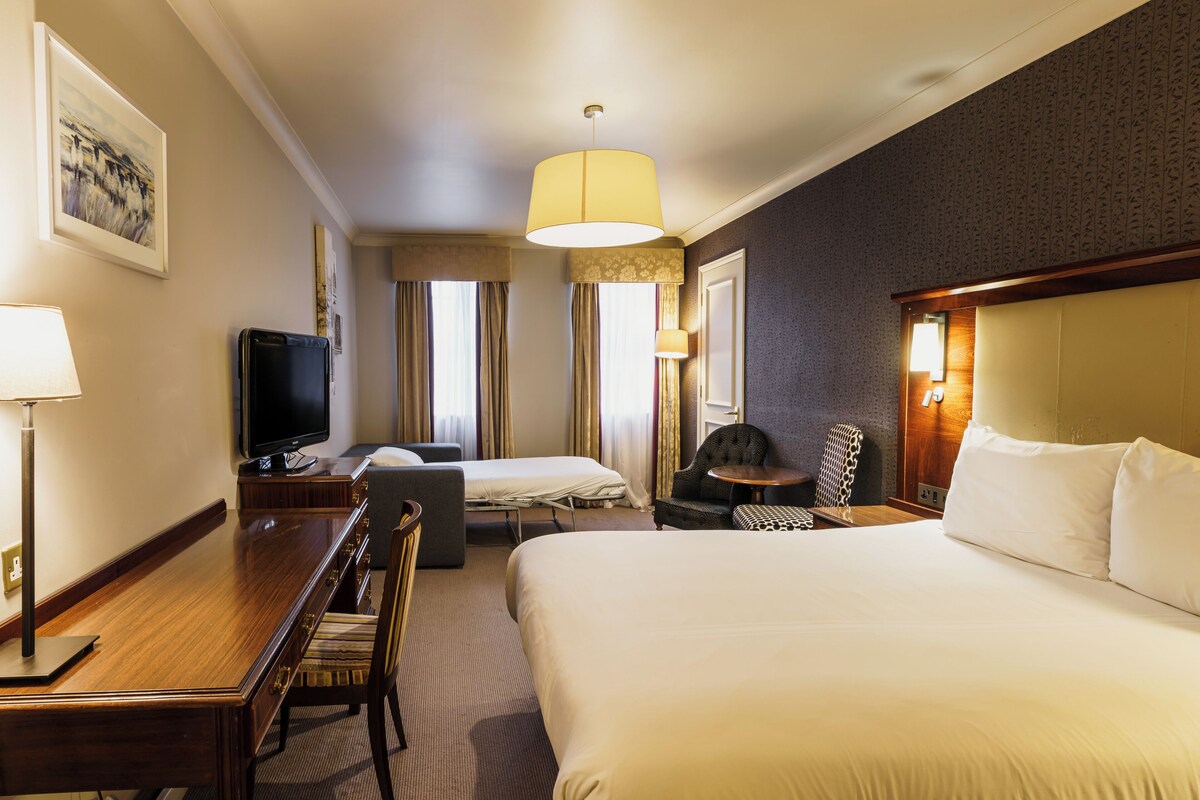 Habitación de hotel con una cama grande, un escritorio con televisor, una silla, una mesita y una lámpara.