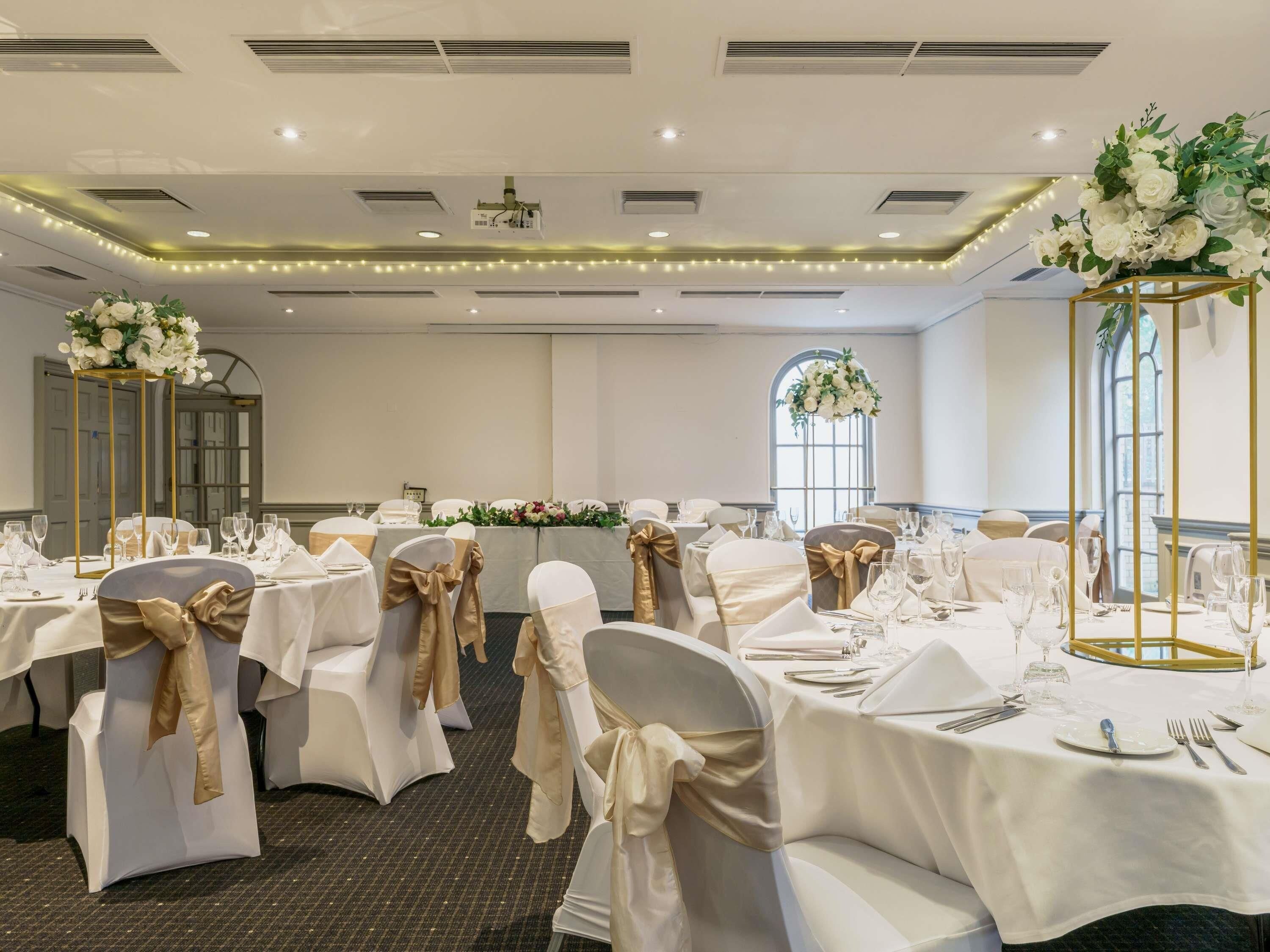 Un salón de banquetes con mesas redondas, manteles blancos y sillas con cintas doradas.