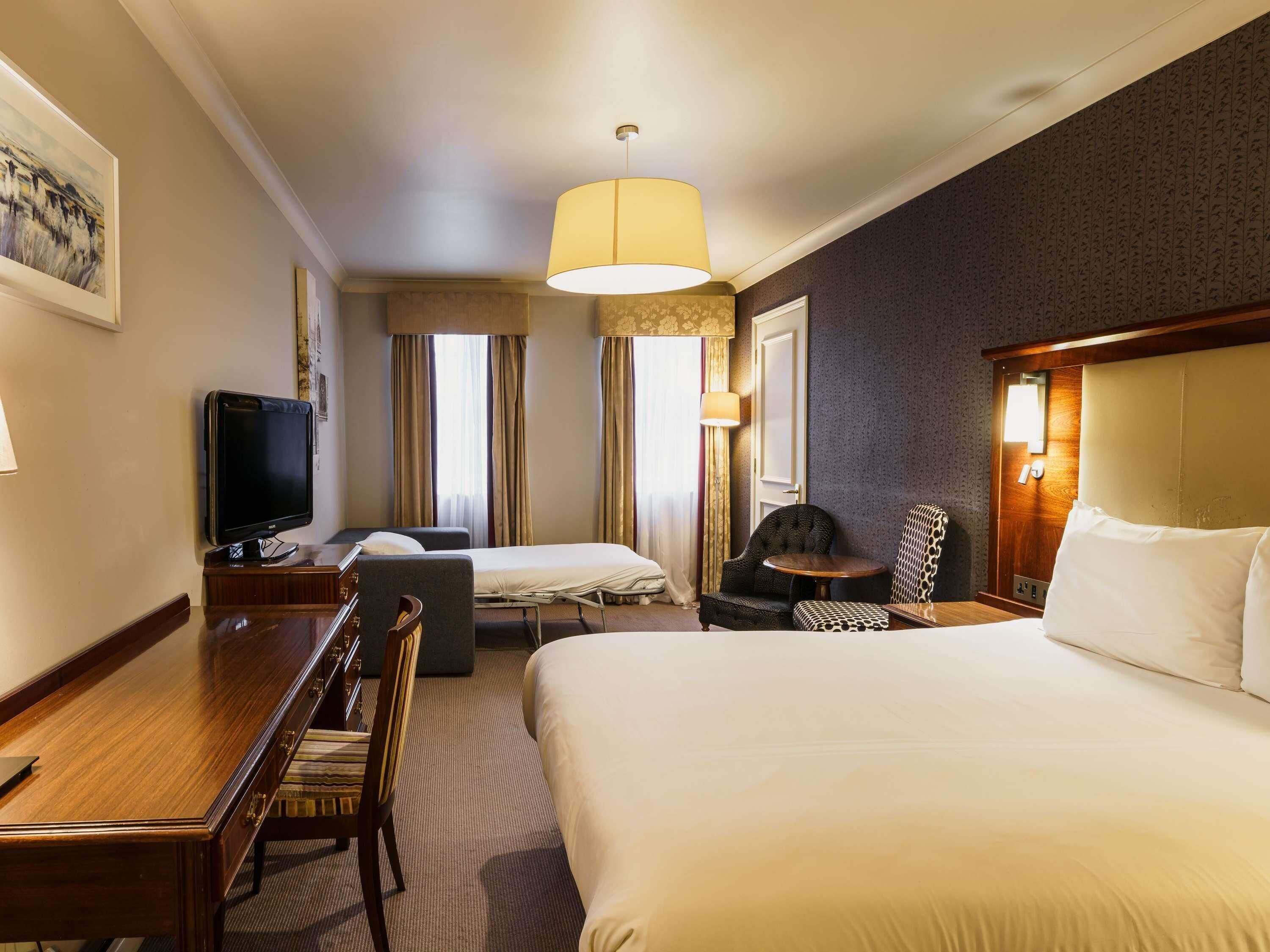 A hotel room with a large bed, a desk with a television, a chair, and a small table.